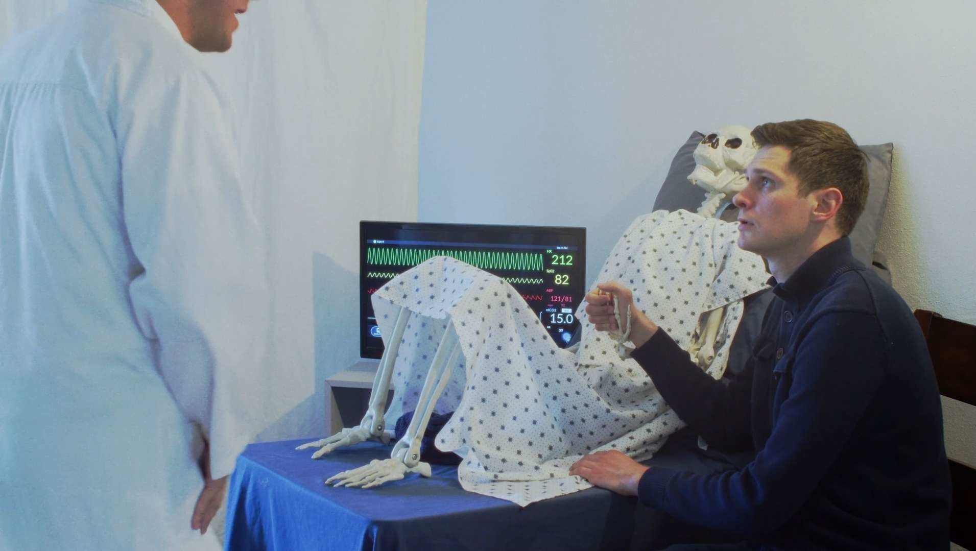 Person in white coat observing a man in a hospital bed with medical equipment and monitors.