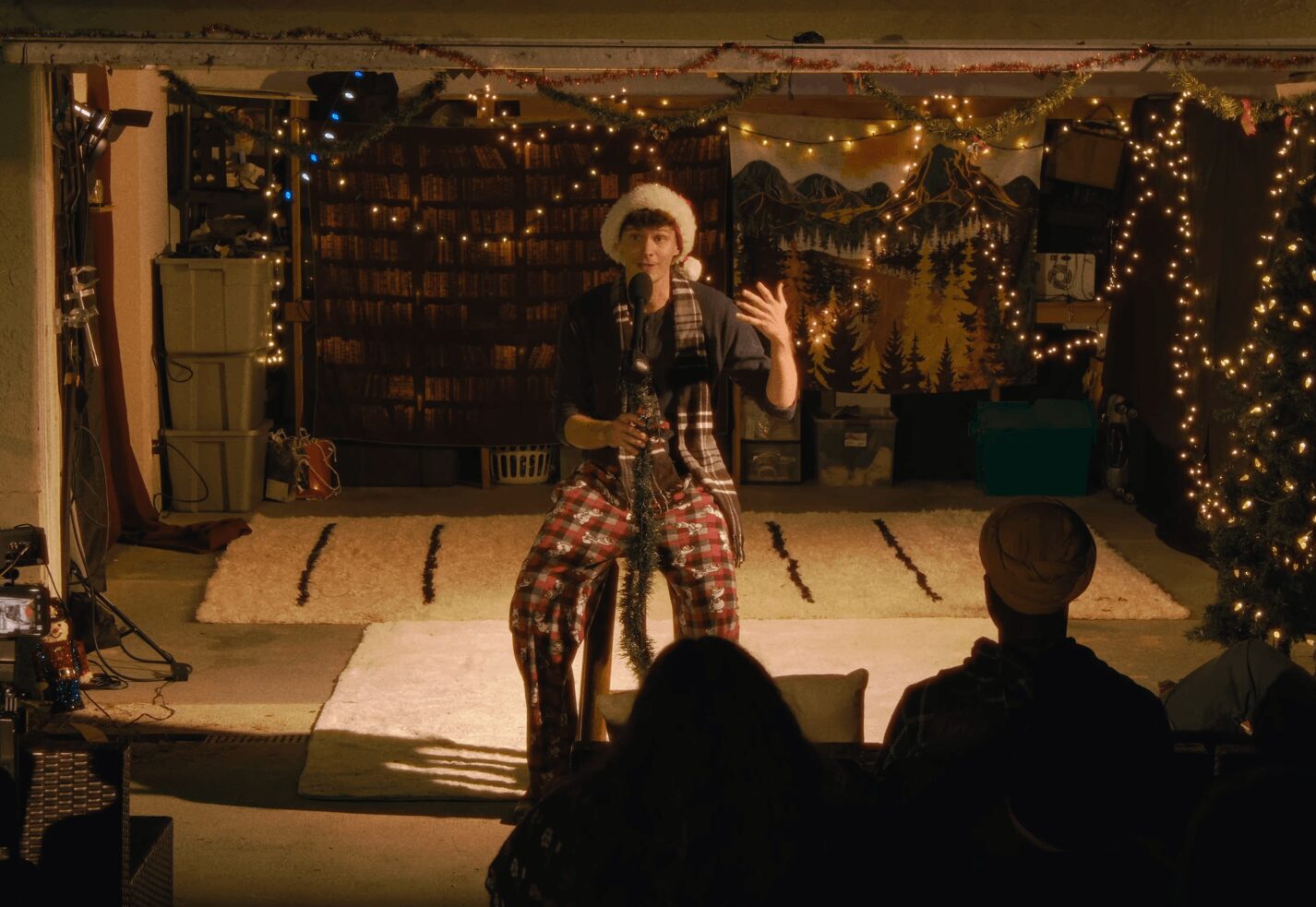 Performer on stage with microphone, wearing hat and patterned pants, in a decorated indoor setting with audience silhouette.
