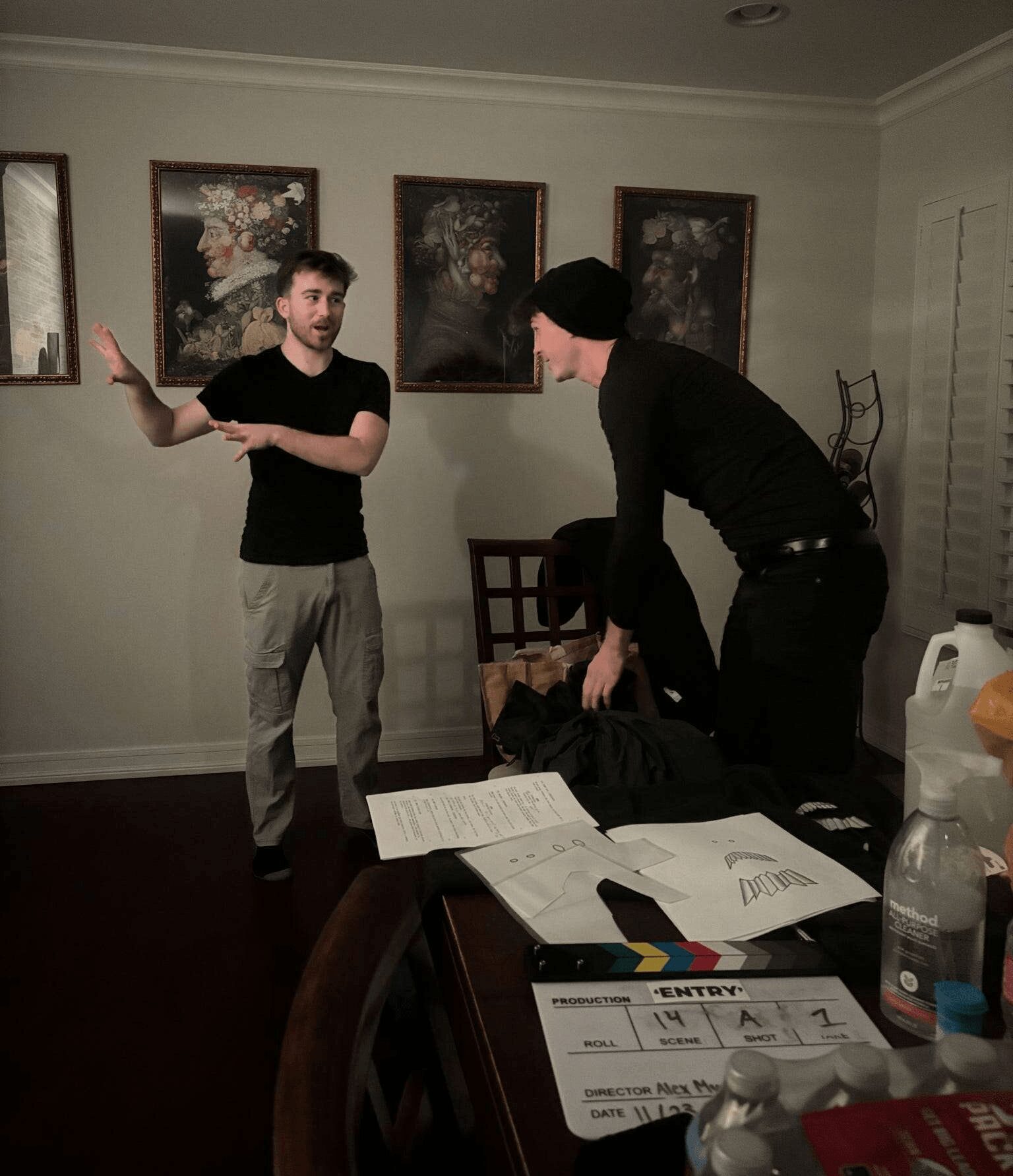Two men in a room with framed artwork, one standing and gesturing, the other leaning over a table with papers and bottles.