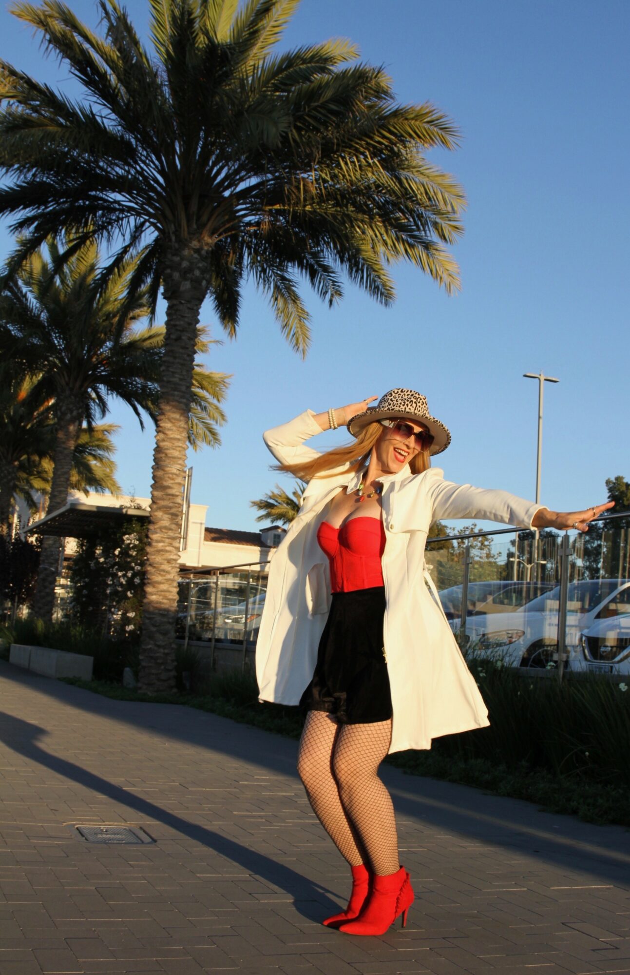 Woman in red top, black shorts, white coat, red boots, and wide-brimmed hat standing outdoors near palm trees and a parking lot.