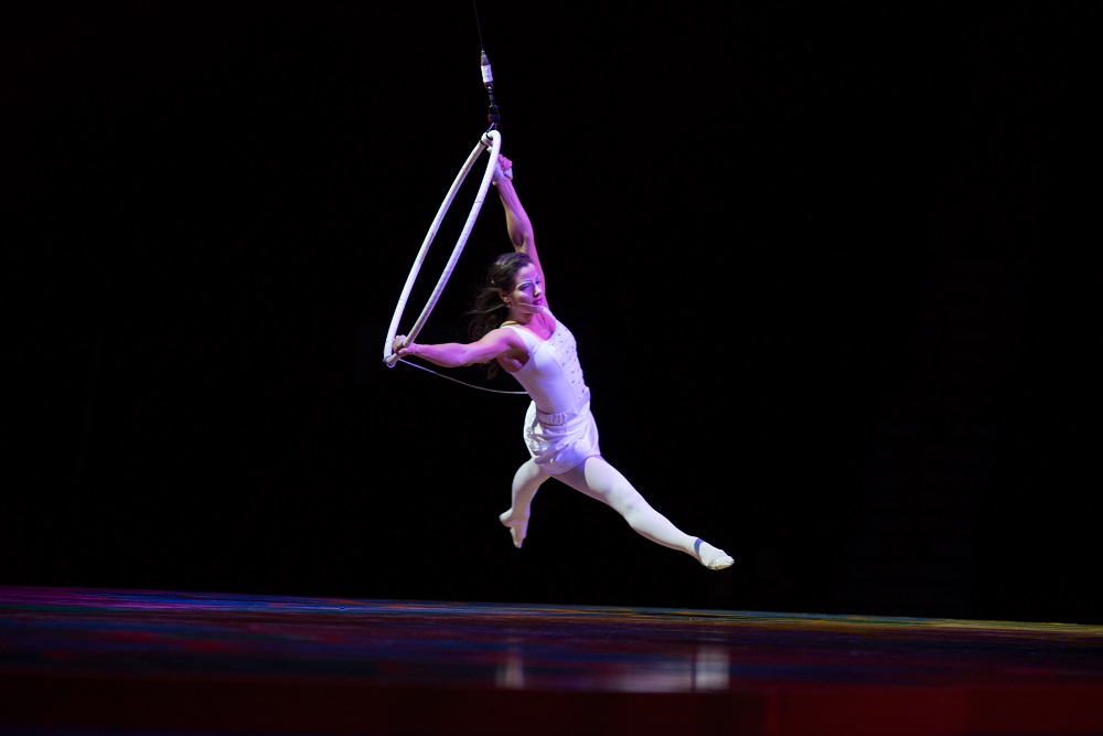 Aerialist performing on a hoop suspended in the air, wearing a white costume, against a dark background.
