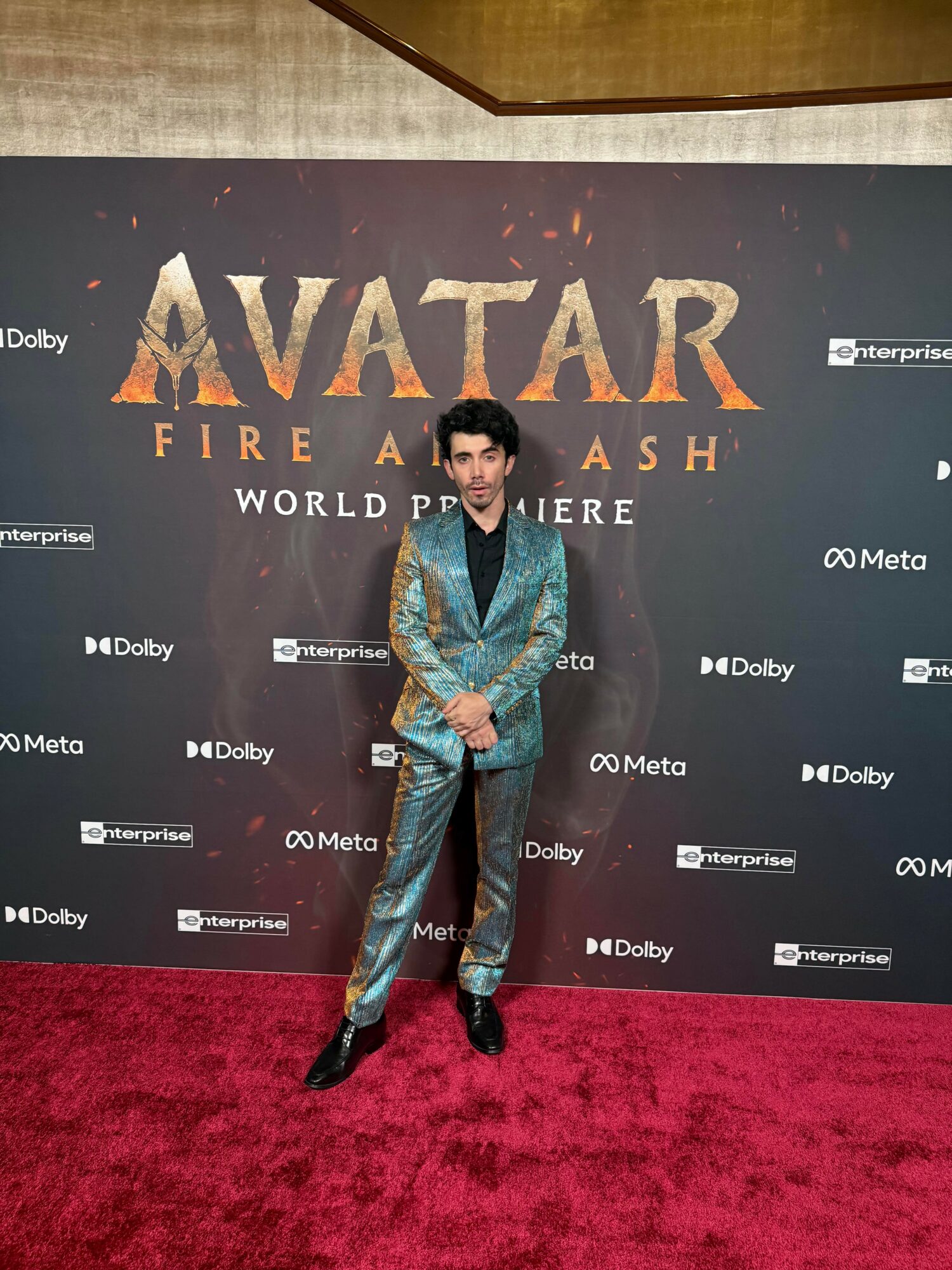 Person in a shiny suit standing on red carpet in front of a promotional backdrop for Avatar: Fire and Ash.