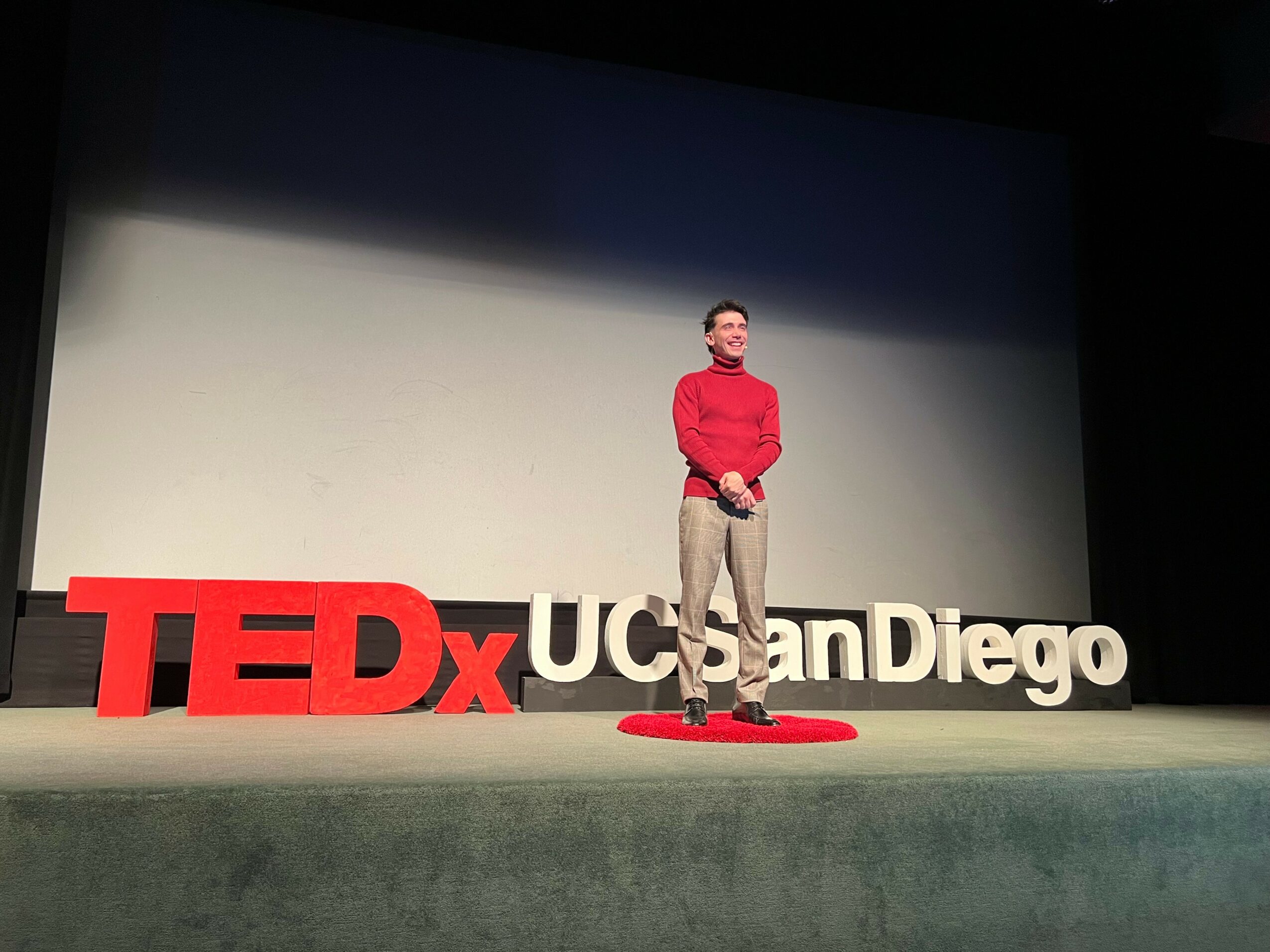Man standing on stage with large TEDxUCSDen Diego sign behind him, wearing a red sweater and beige pants.