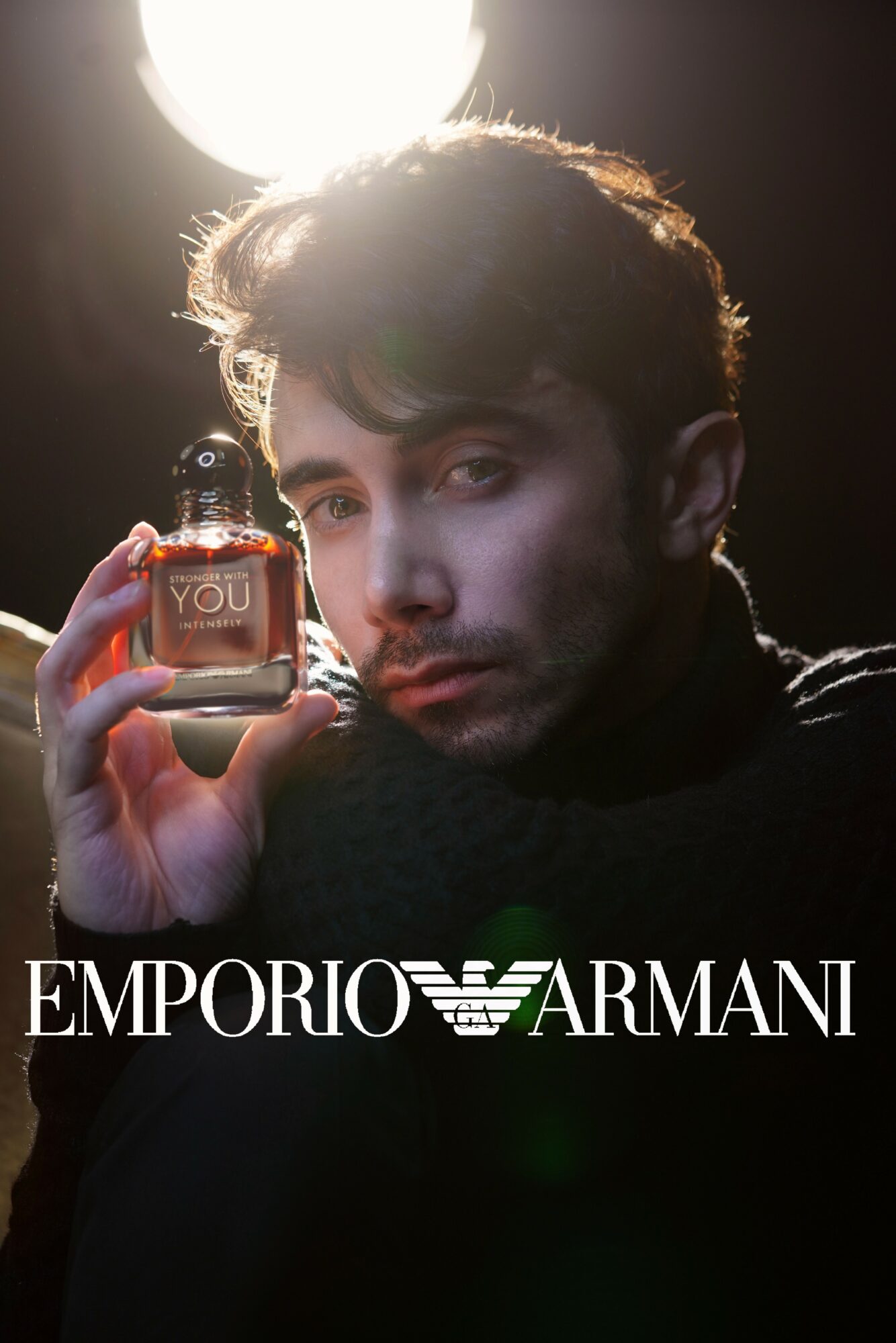 Young man with dark hair holds a small perfume bottle in front of a bright light, with a dark background.