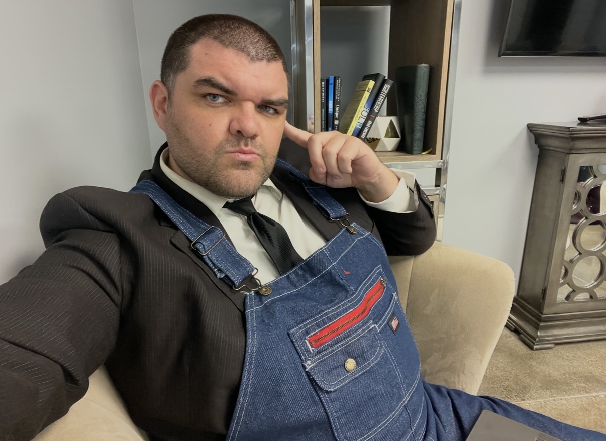 Man in suit and apron sitting on a beige chair, adjusting his collar, in a room with bookshelves and a TV.