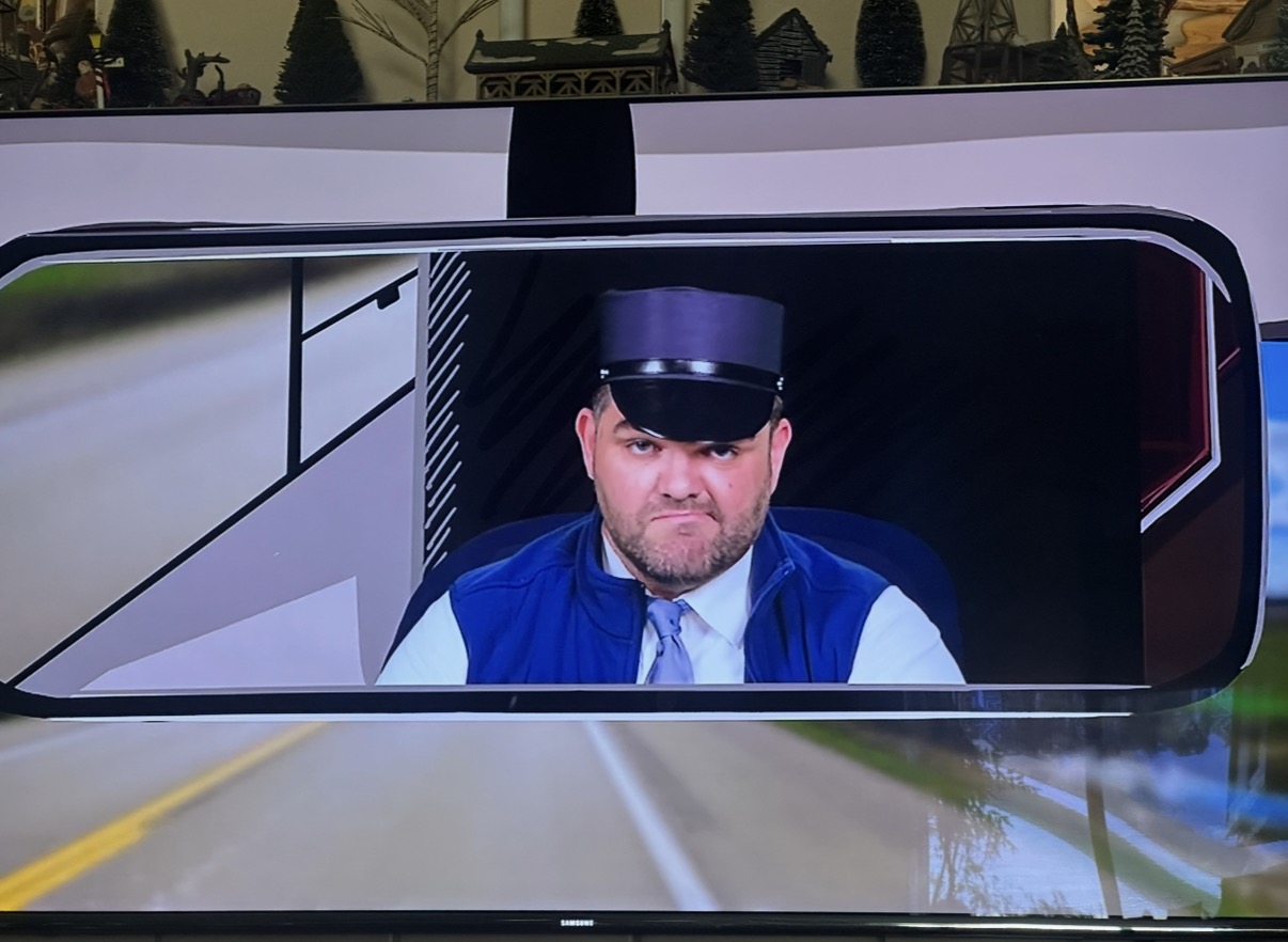 Person wearing a uniform and cap, seen through a vehicle's rearview mirror, with a road and trees in background.