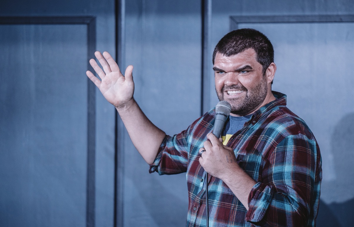 Man with dark hair and beard speaking into a microphone, smiling, wearing a plaid shirt, gesturing with his hand.
