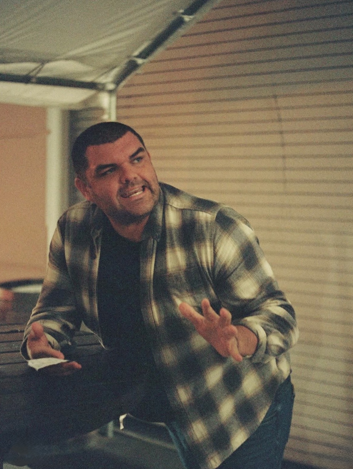 Man smiling and gesturing with hand, wearing plaid shirt indoors with closed blinds behind him.