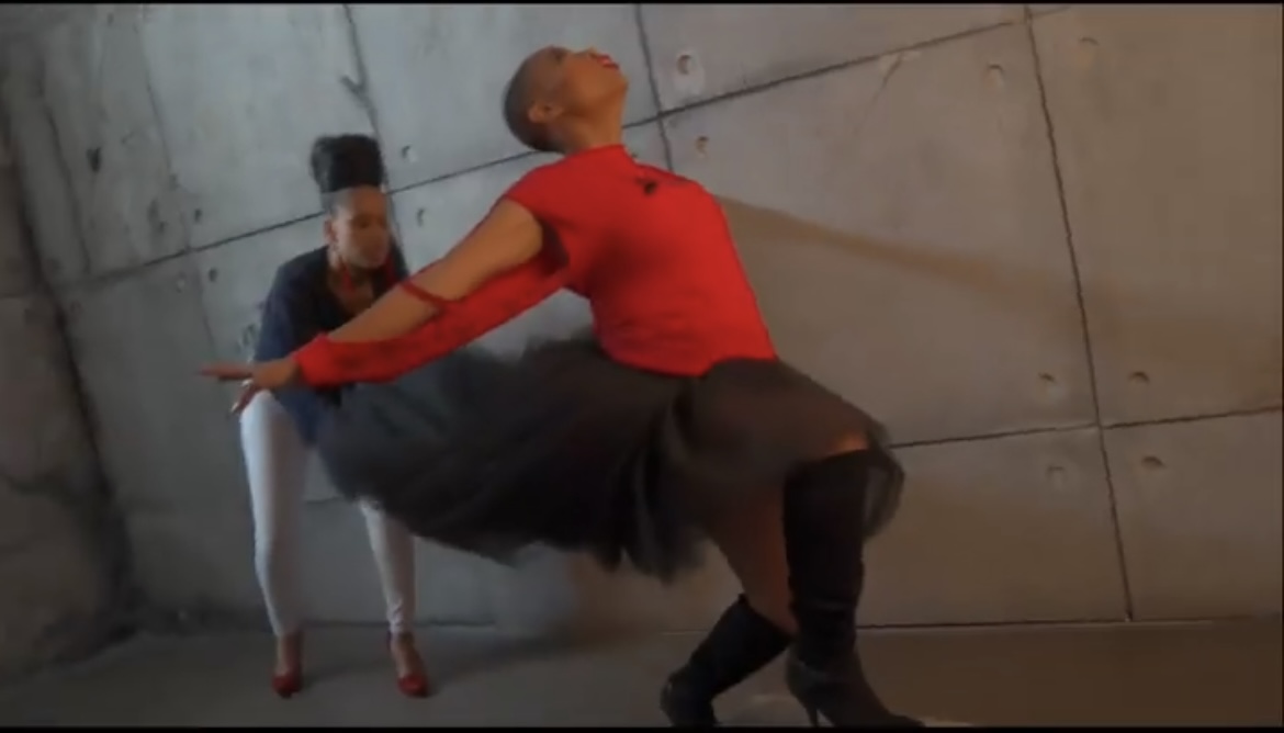Two dancers perform in front of a concrete wall, one in a red top and black skirt, the other in black and white.