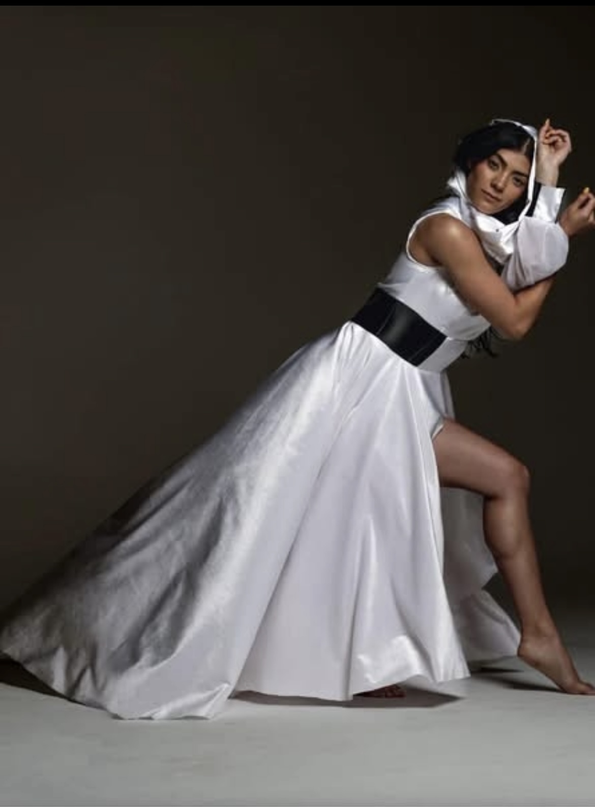 Woman in a white gown with black belt, posing with one leg raised, against dark background.