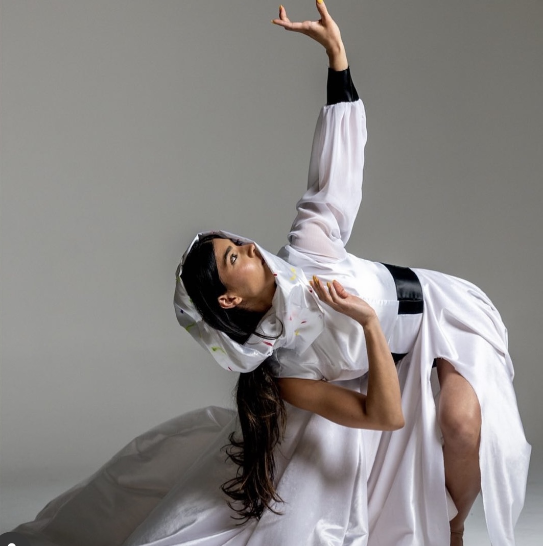 Person in a white costume with a hood, posing with one arm raised and the other near the chest.