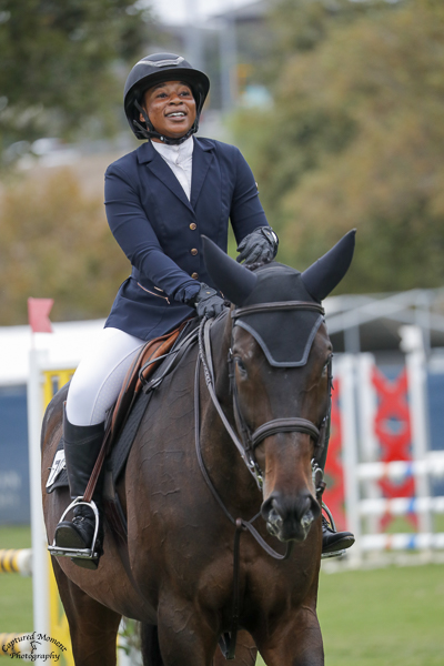 Person riding a horse outdoors, wearing a helmet, navy jacket, white pants, and gloves, smiling with trees and show jumps in background.