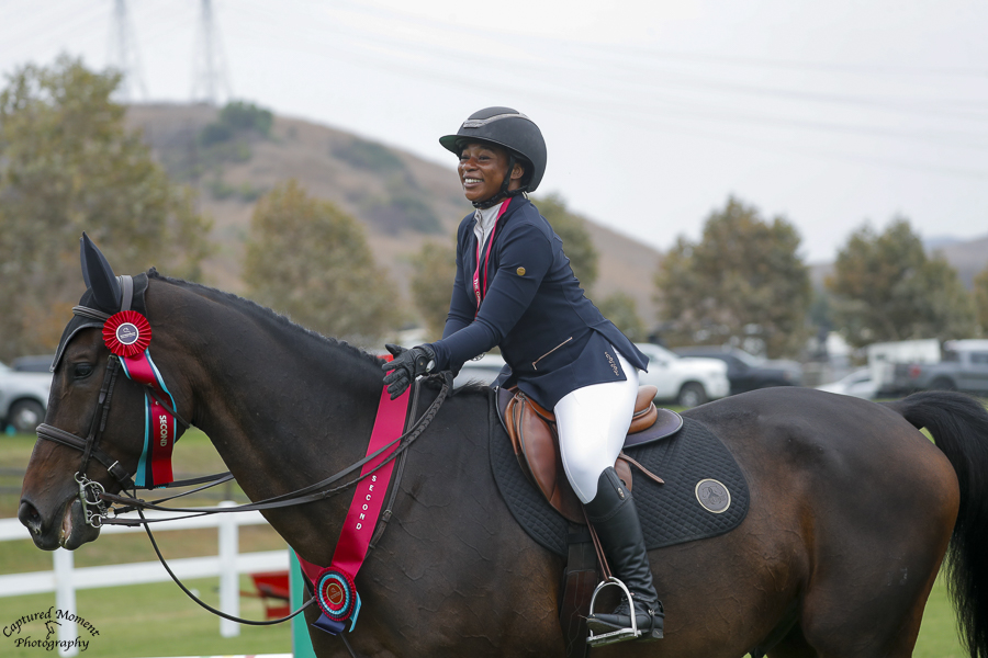 Person riding a dark horse with a red ribbon on its bridle outdoors, wearing a helmet and riding attire.