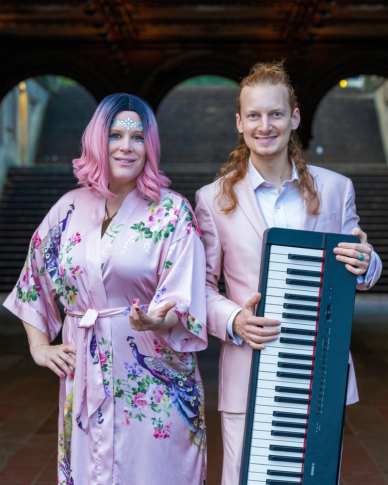 Two women standing outdoors, one holding a keyboard, smiling, with a bridge in the background.