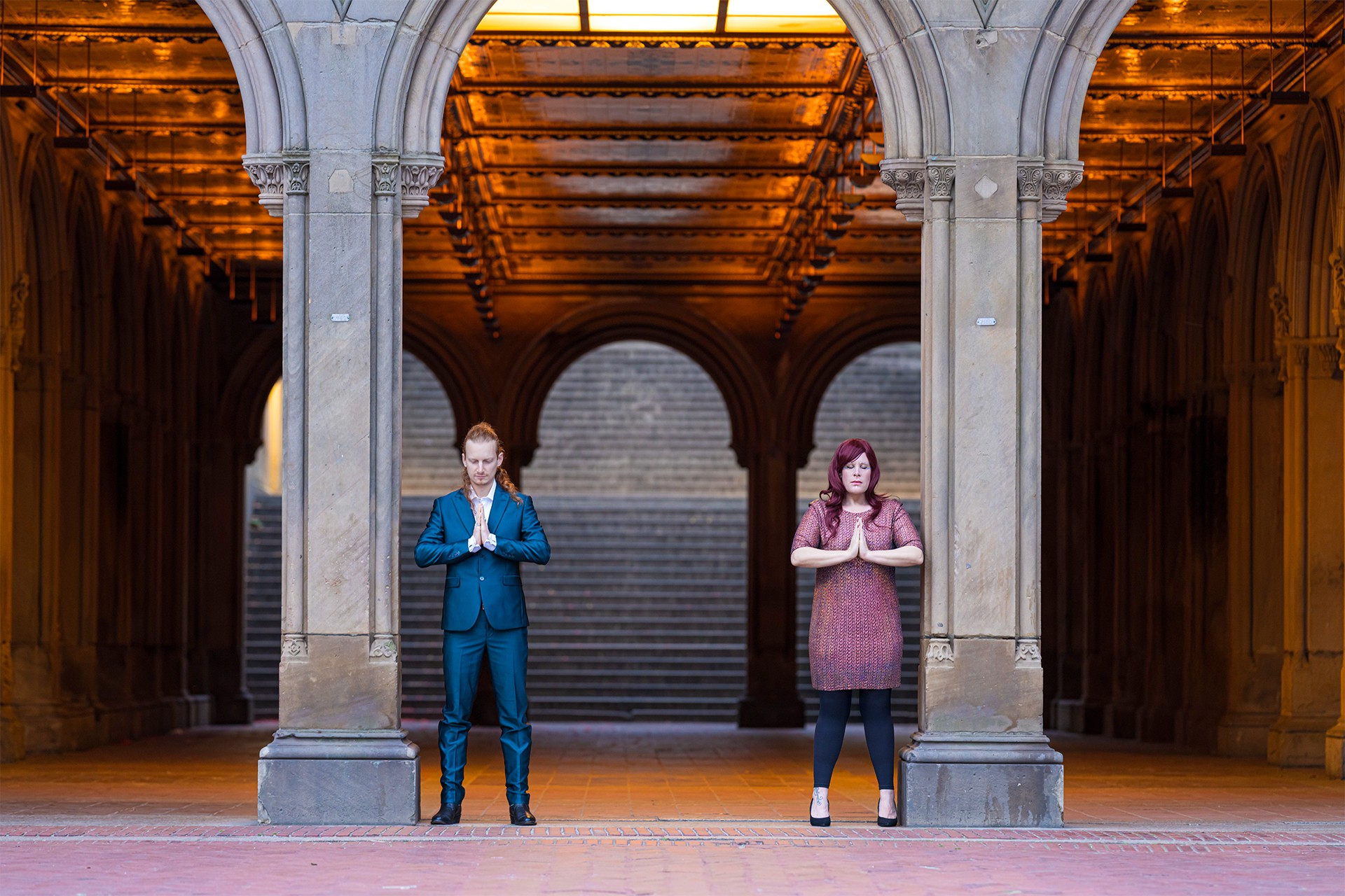 Two people standing under an ornate archway with stairs in the background, one in a blue suit and the other in a pink dress.