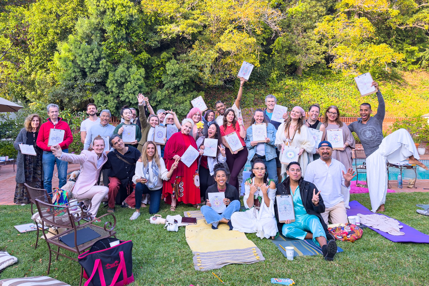 Group of people outdoors on grass, some holding papers, with trees in background, sitting and standing near blankets and chairs.