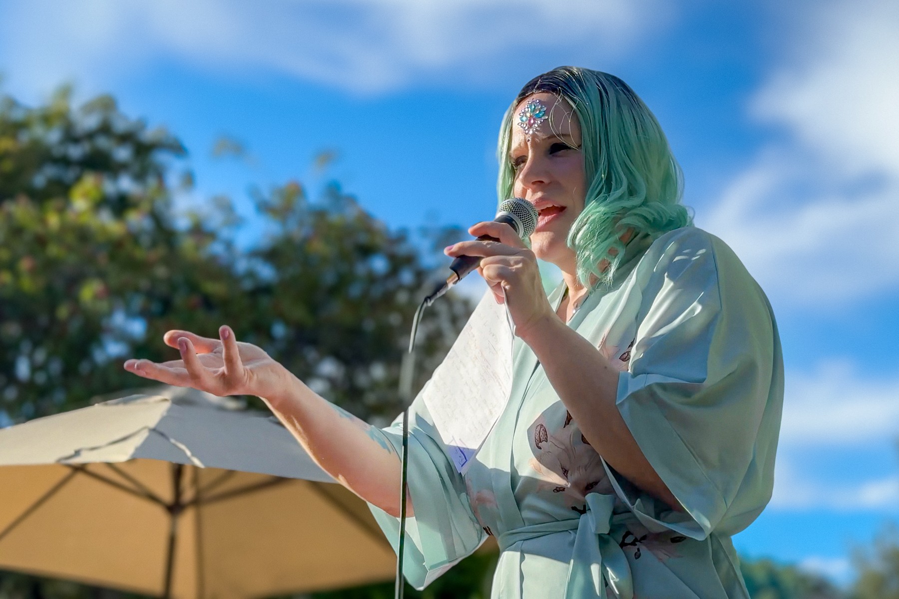 Person with teal hair speaking into a microphone outdoors under a blue sky with clouds and trees.