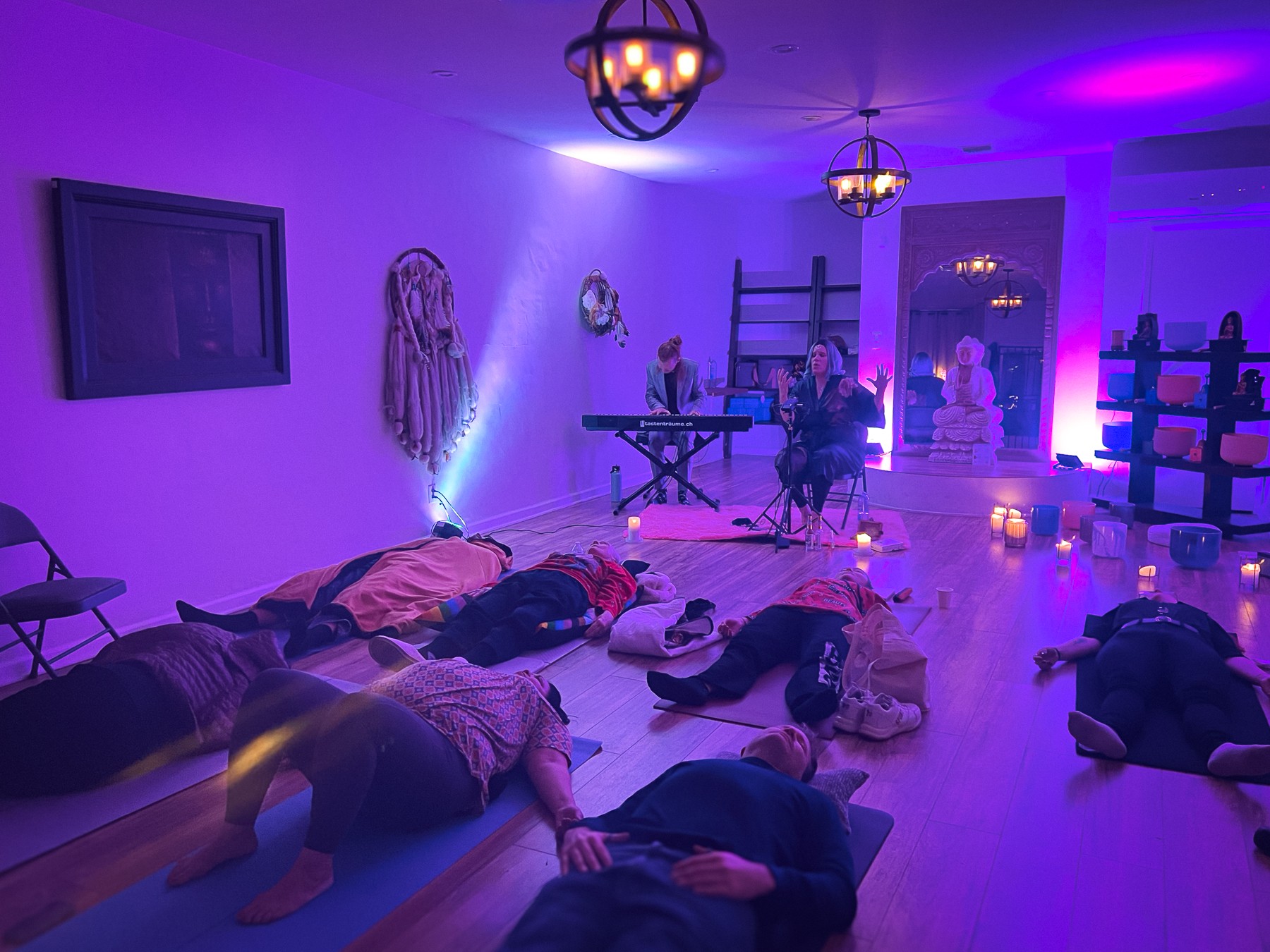 People lying on the floor in a dimly lit room with purple and blue lighting, some with arms outstretched, others resting.