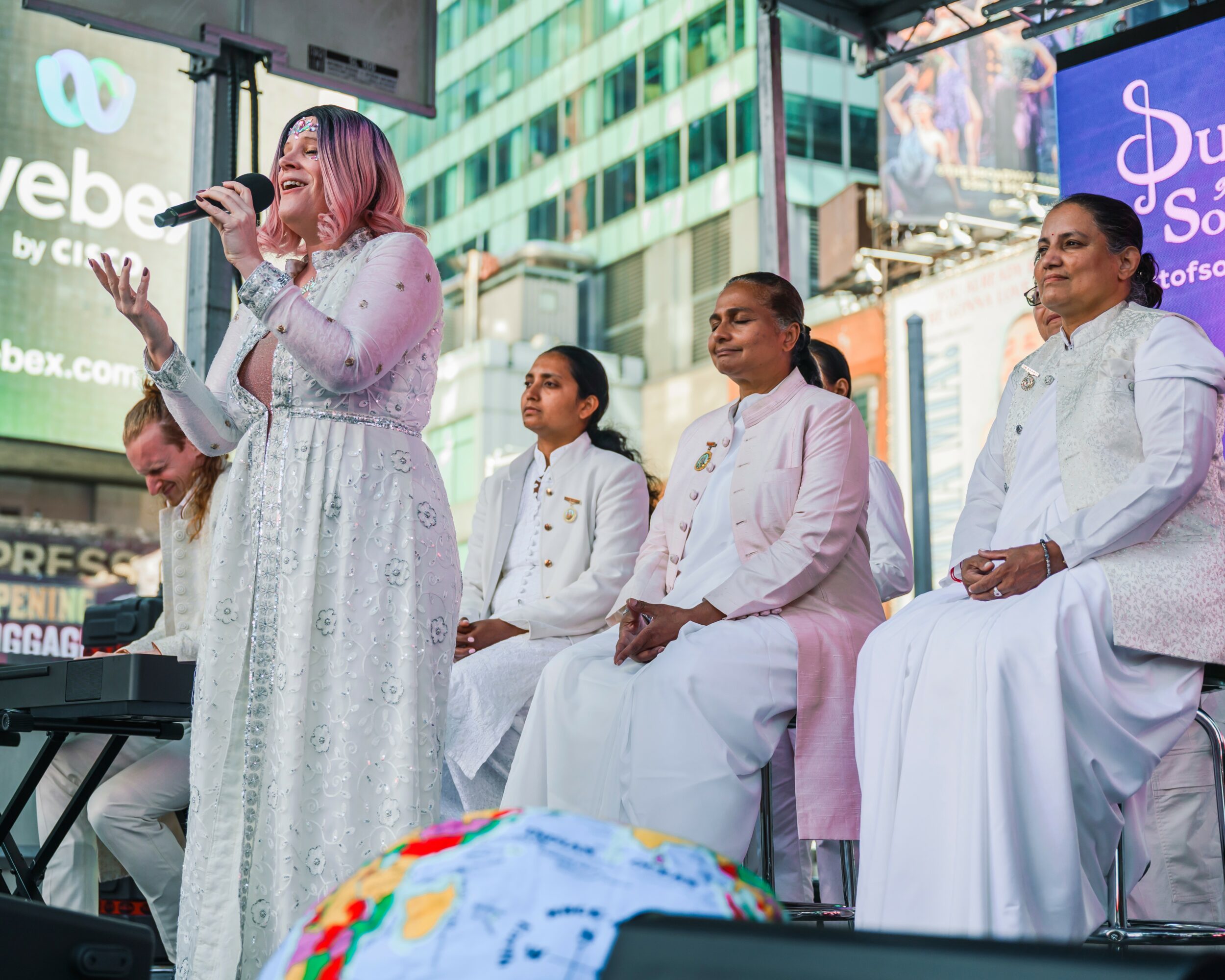 Four women seated on stage, one woman standing and speaking into a microphone, city buildings in background.