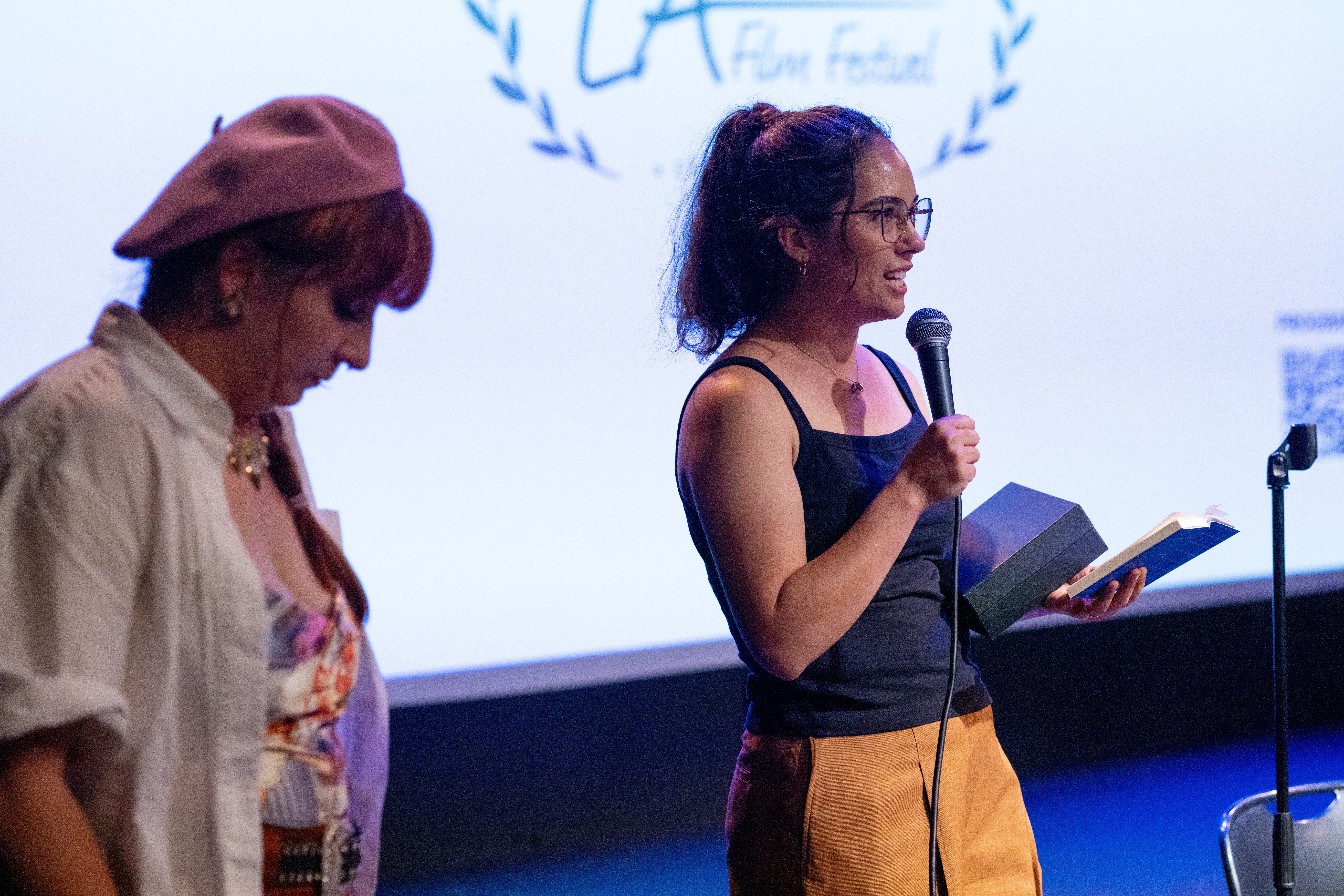 Two women on stage, one speaking into a microphone, the other looking down, with a large screen behind them displaying a logo.
