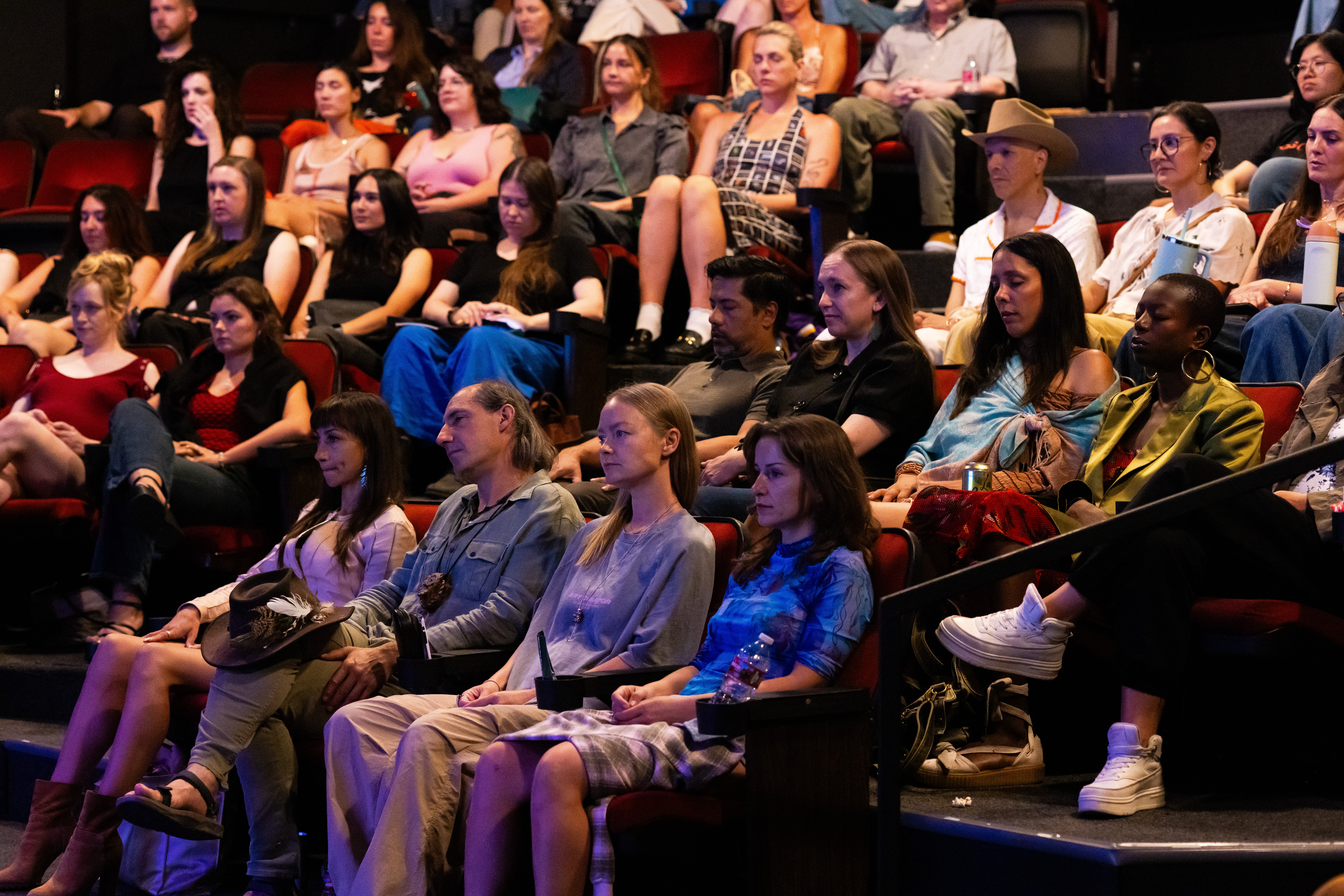 Audience seated in theater, watching a performance or presentation, with diverse individuals in casual attire.