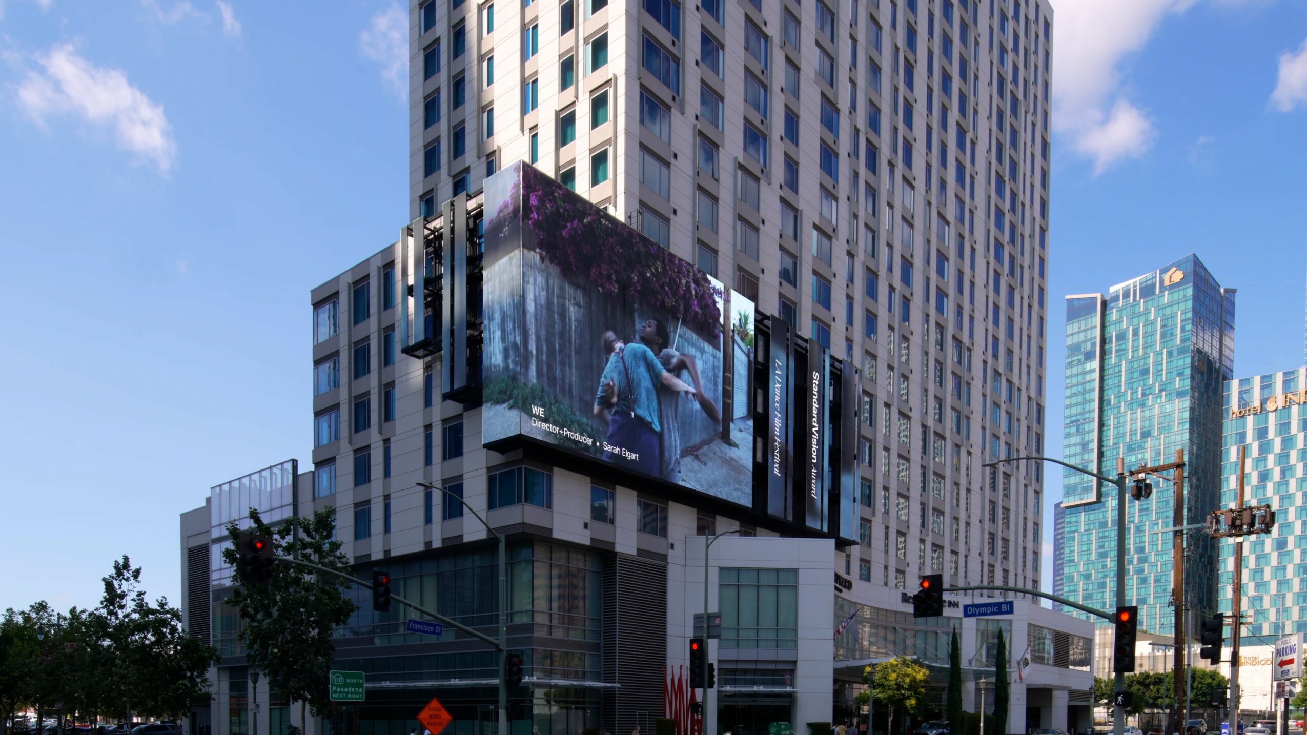 Tall building with digital billboard displaying a person in a blue jacket, surrounded by other skyscrapers and a street scene.