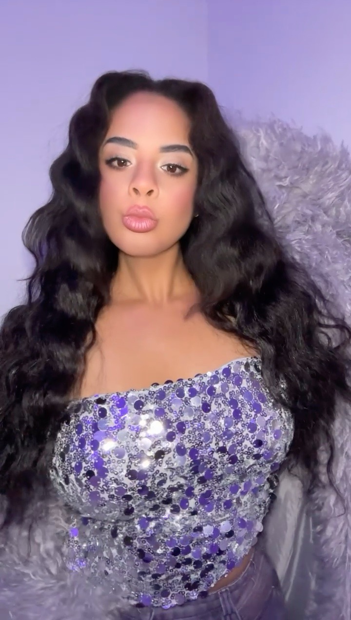 Young woman with long wavy hair wearing a sparkly purple top, looking at the camera.