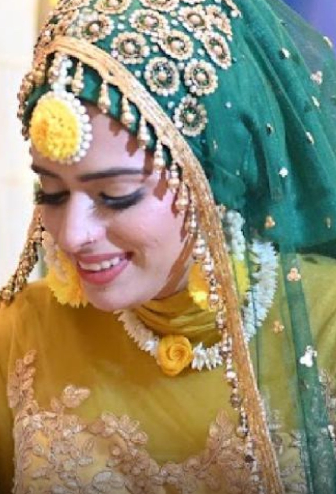 Woman wearing traditional attire with a green headscarf and jewelry, smiling with eyes closed.