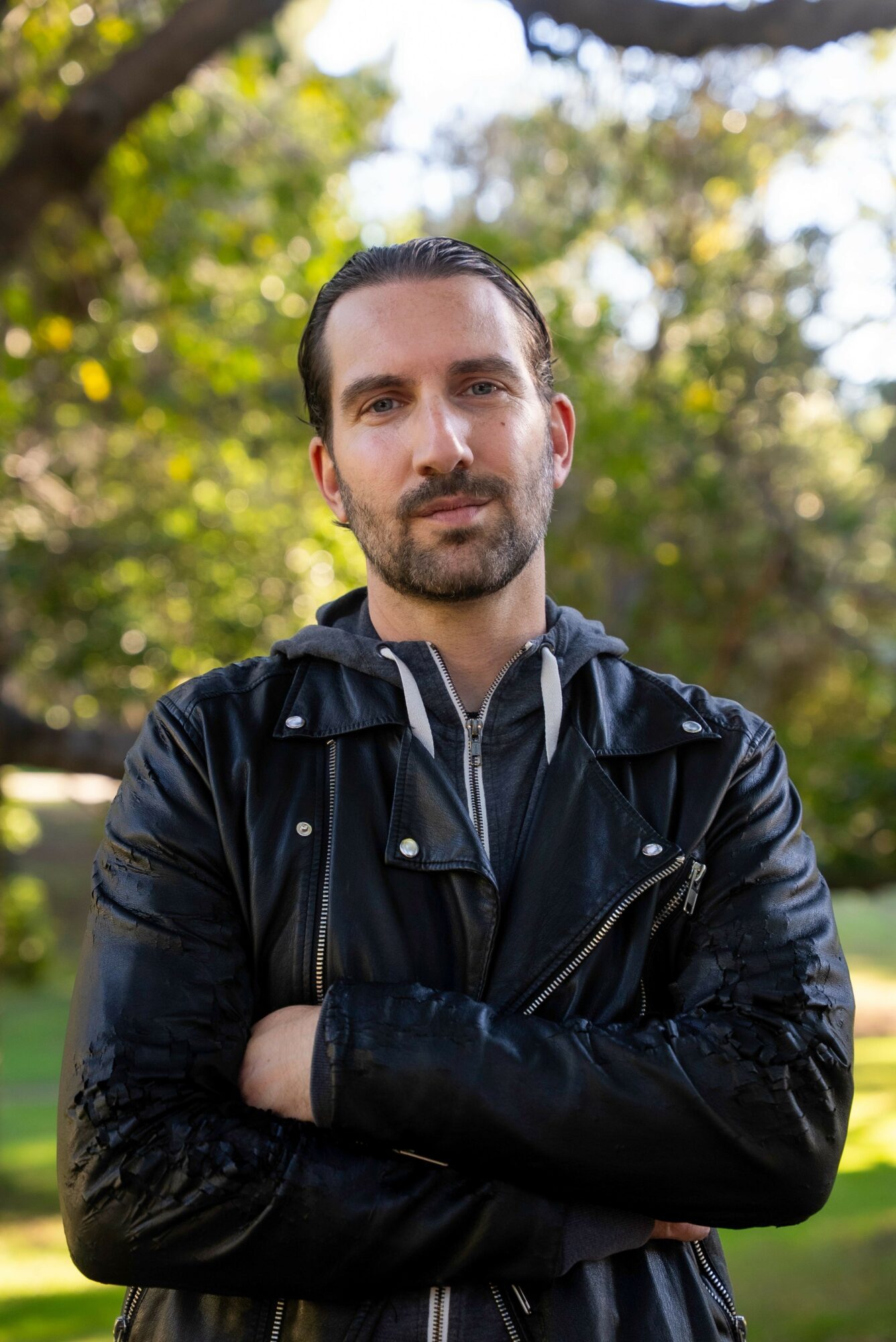 Man with dark hair and beard wearing a black jacket outdoors with trees and sunlight behind him.