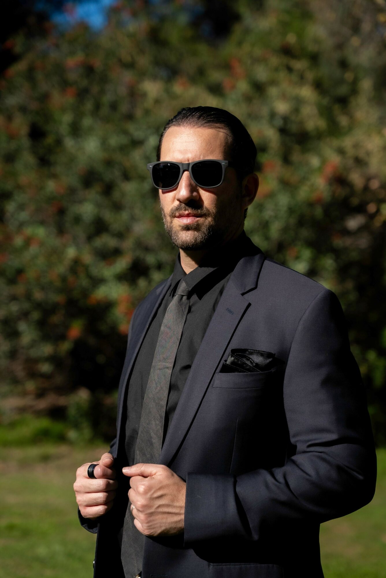 Man in sunglasses and suit standing outdoors with trees in background.