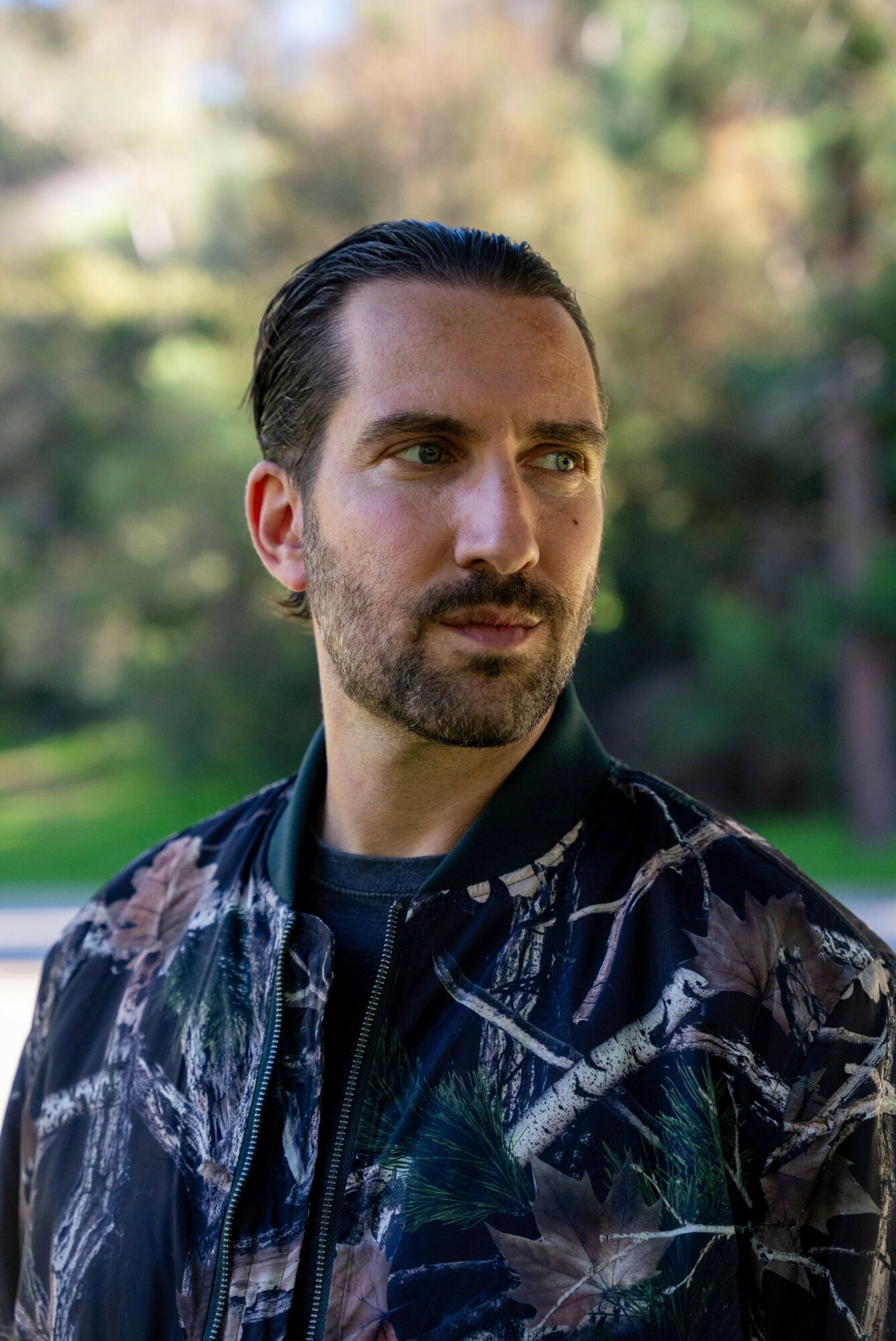 Man with dark hair and beard outdoors, wearing a patterned jacket, looking to the side.