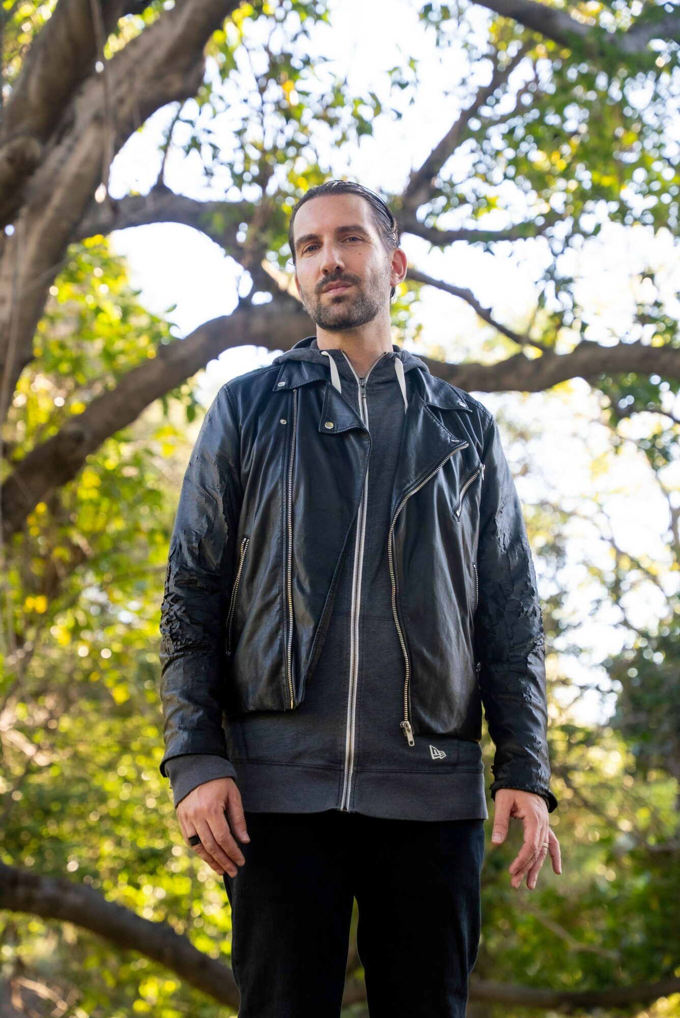 Man standing outdoors near a large tree with green leaves, wearing a black jacket and dark pants.