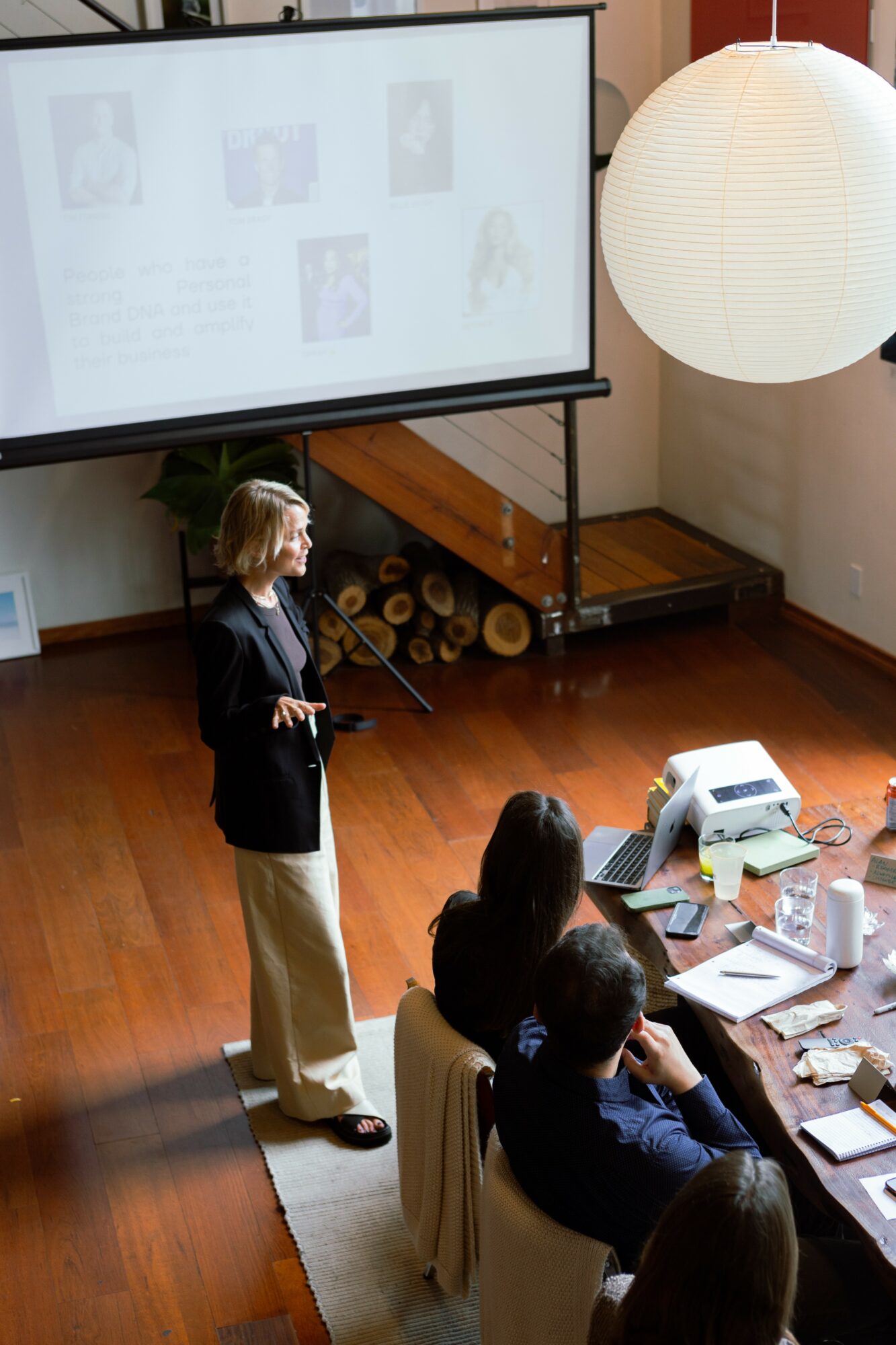 Woman presenting to seated audience in a room with a large screen and a round paper lantern ceiling light.