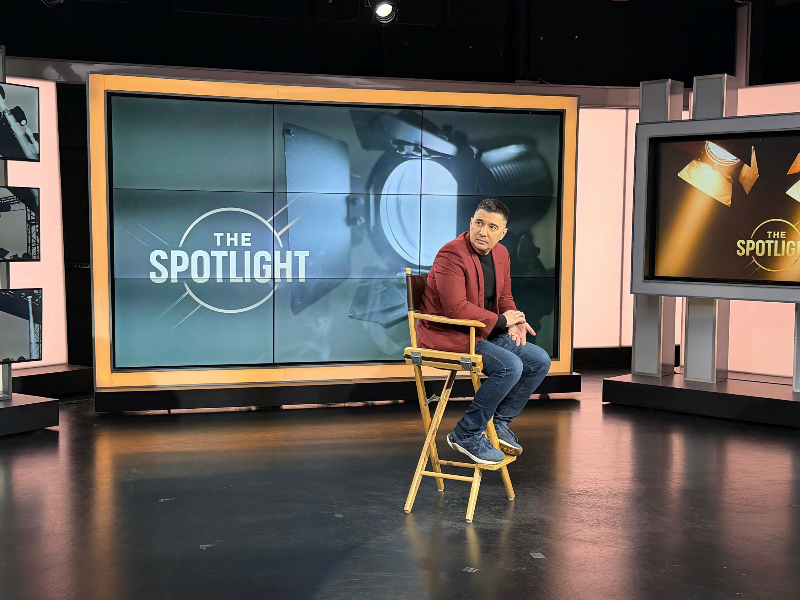 Man sitting on a wooden chair in a television studio with large screens displaying 'The Spotlight' behind him.