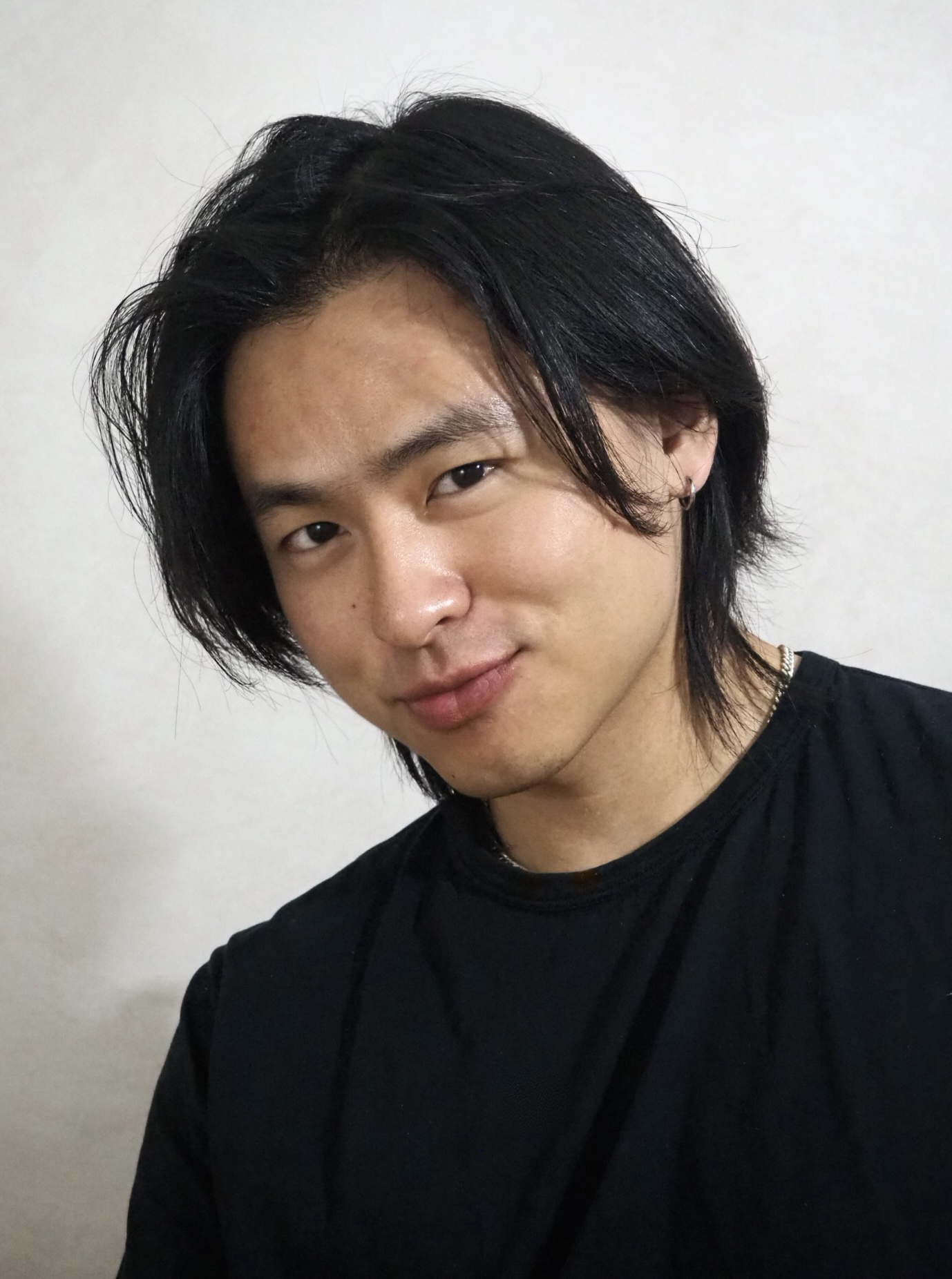 Young man with black hair and a slight smile, wearing a black shirt, against a plain background.