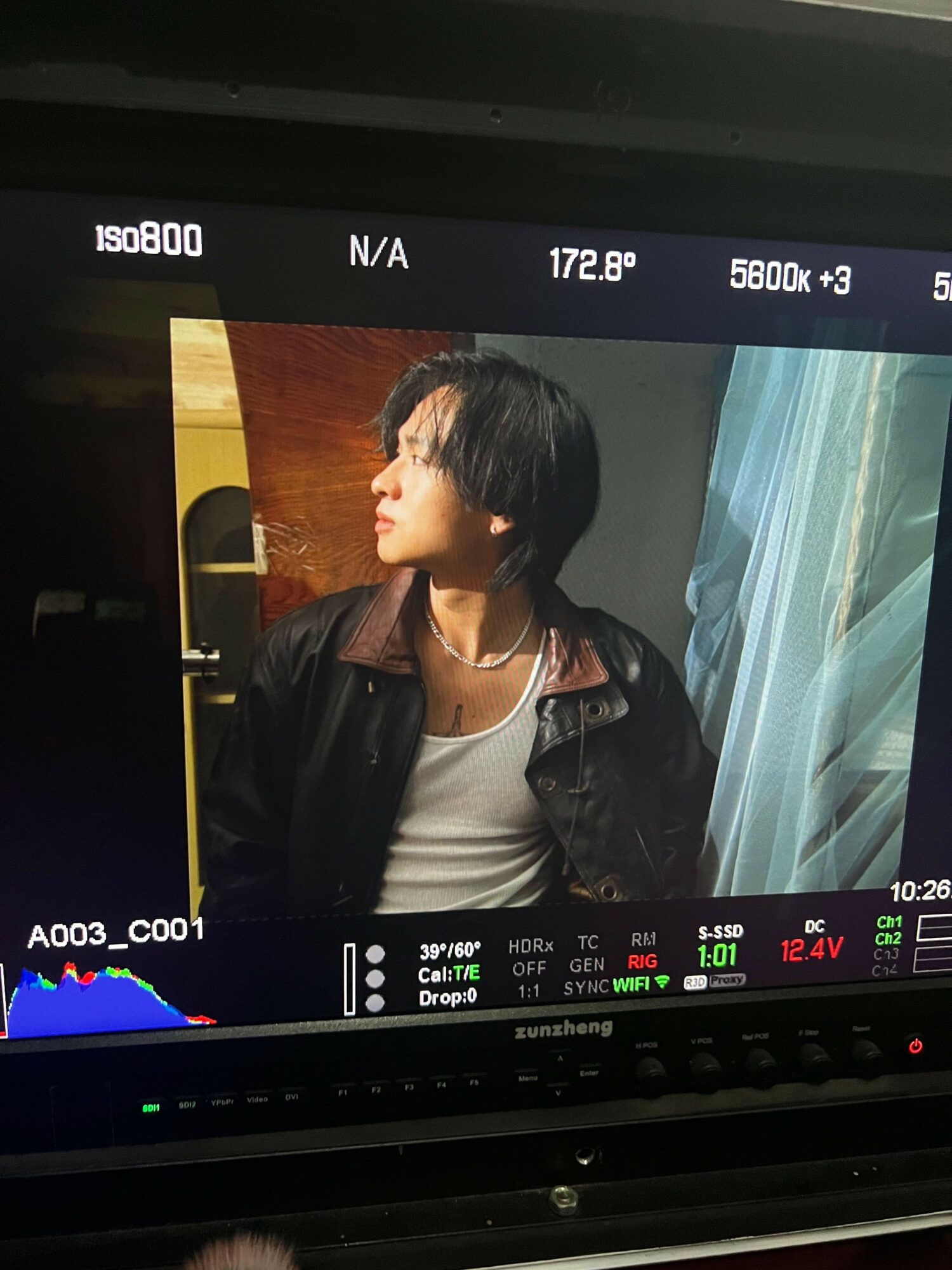 Person with short dark hair wearing a black jacket and white shirt, looking to the side, standing near a window with curtains.