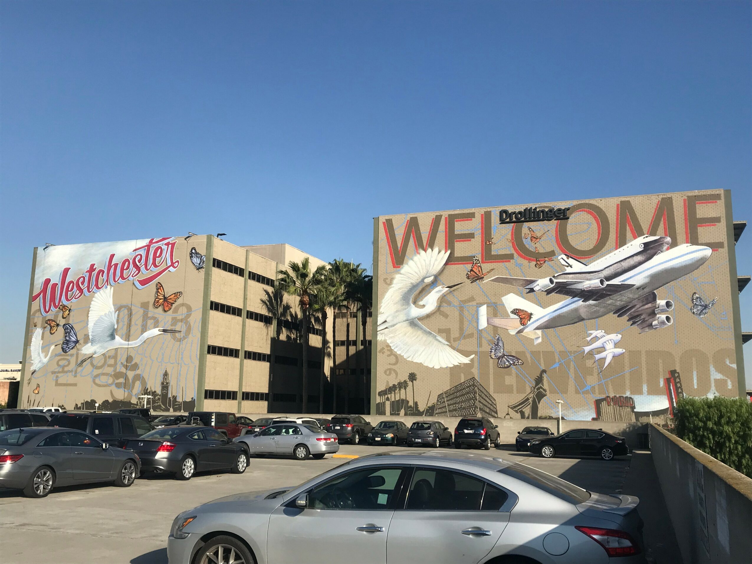 Large building with murals, cars parked in front, clear blue sky, and palm trees nearby.