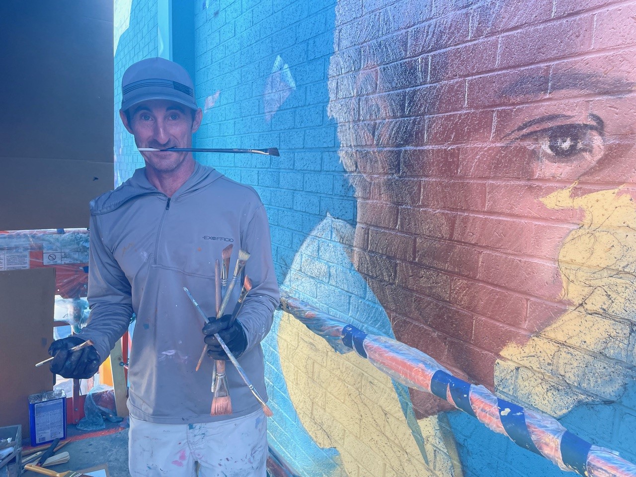 Person wearing a hat and gloves standing next to a colorful mural on a brick wall.