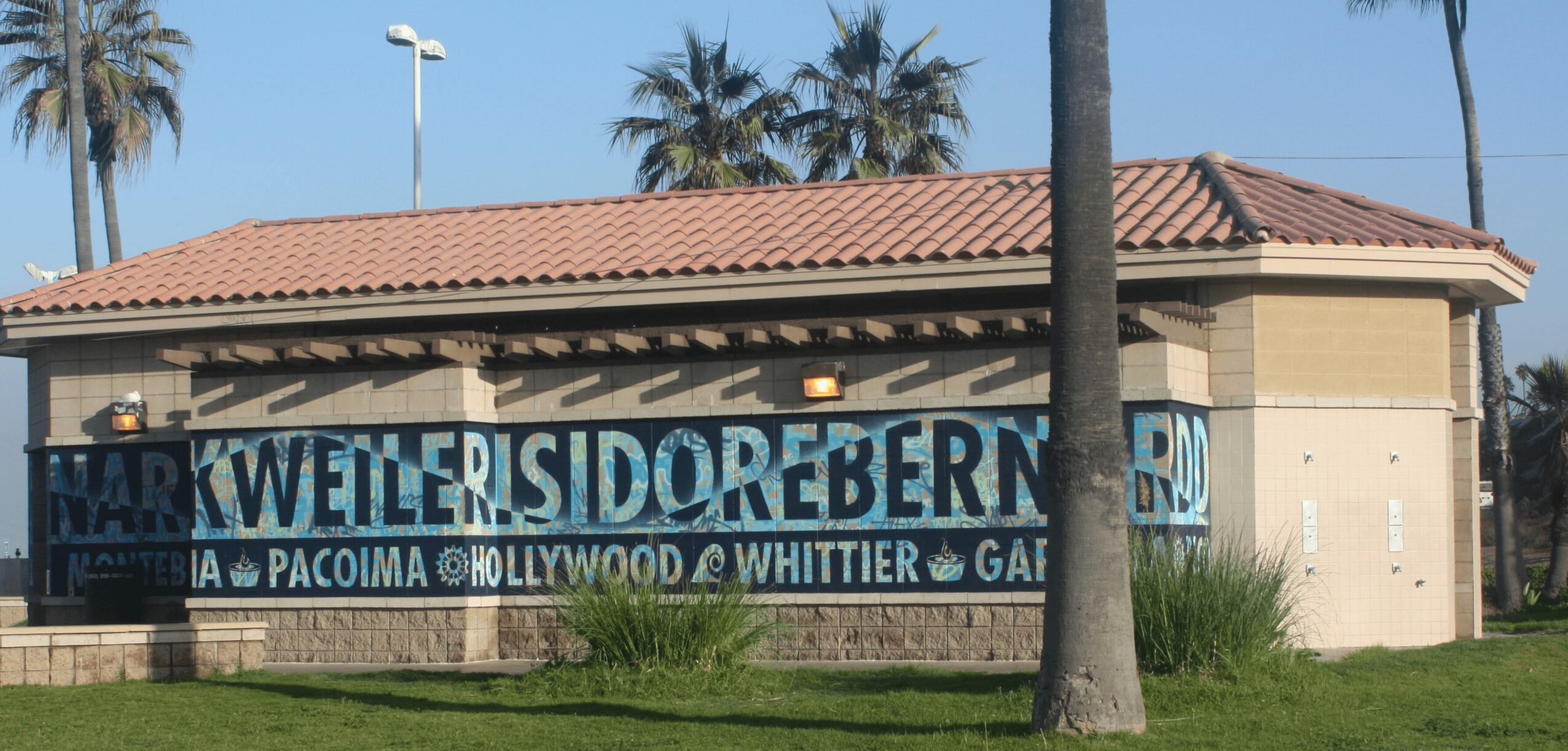 Building with a large banner displaying various names and words, surrounded by palm trees and a grassy area.