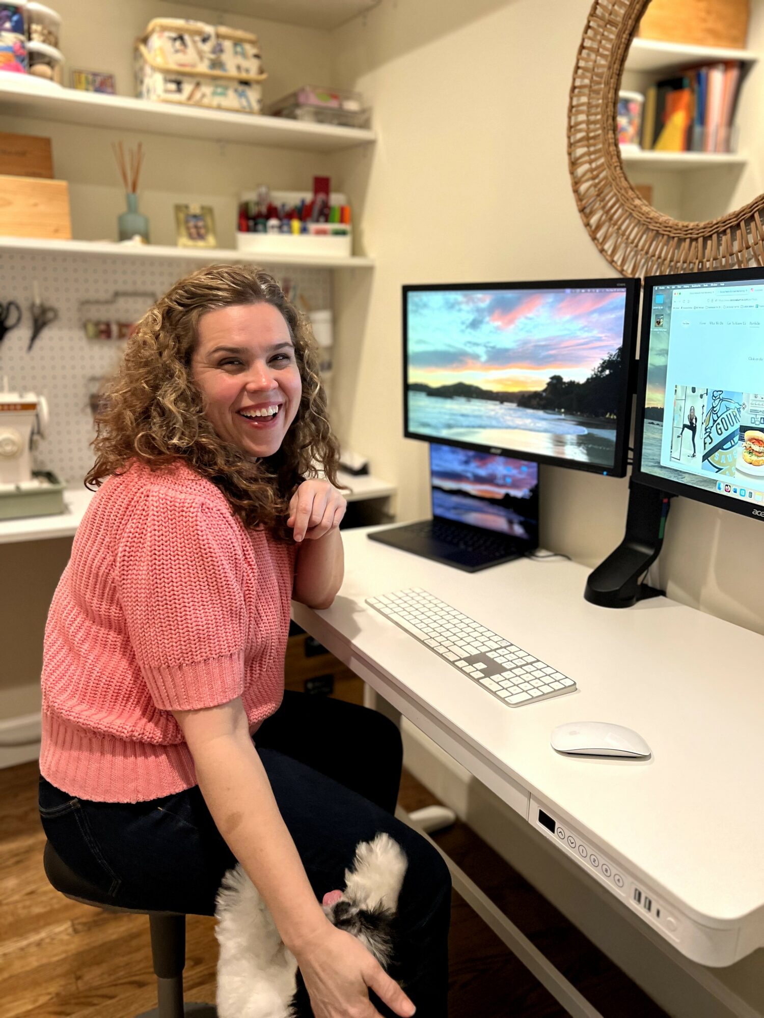 Smiling woman with curly hair sitting at a desk with two monitors, in a room with shelves and a mirror.