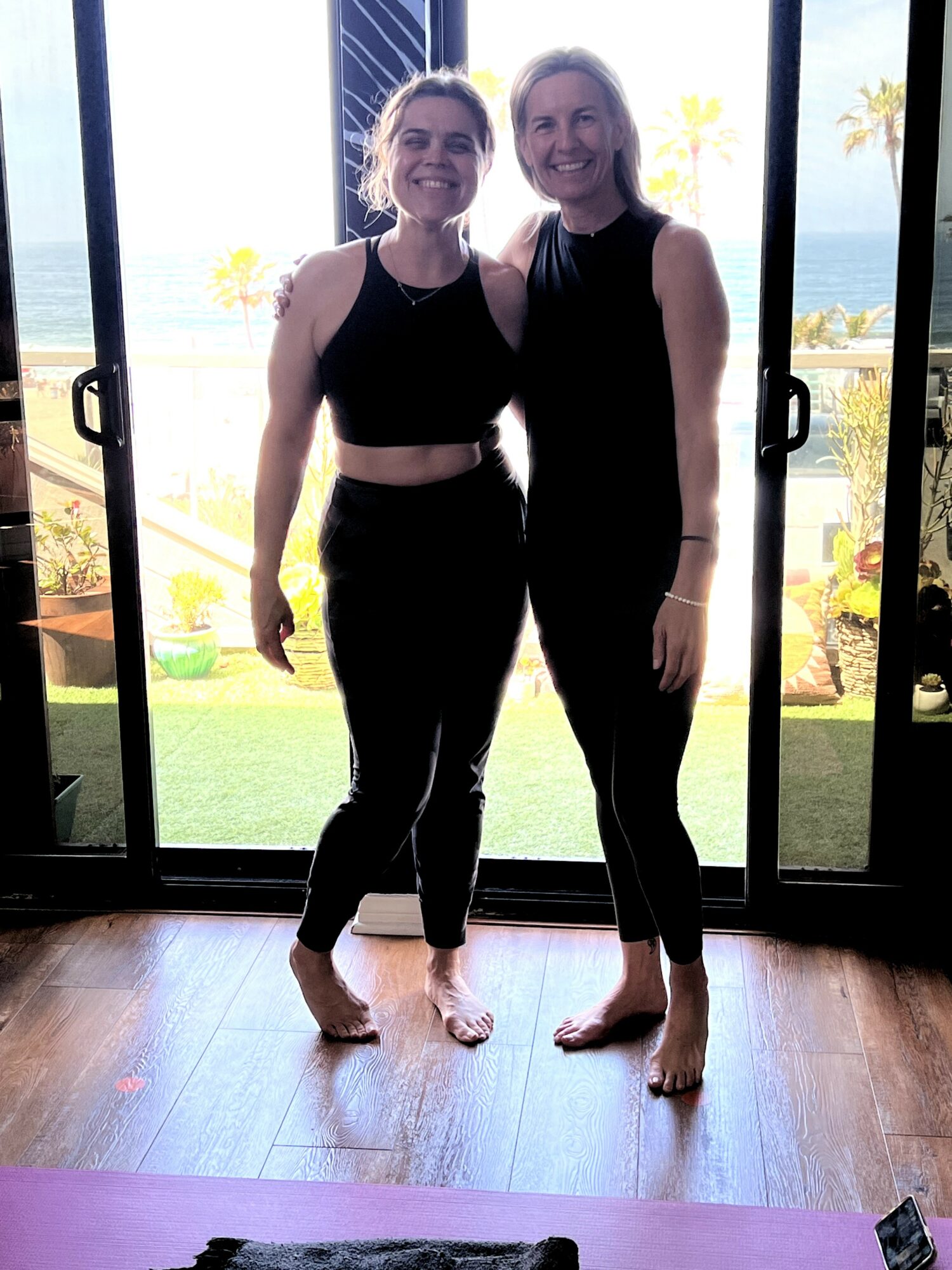 Two women standing barefoot indoors, smiling, with a view of a garden and ocean through glass doors behind them.
