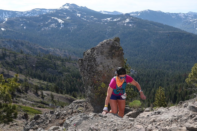 Person hiking on rocky trail in mountainous landscape with trees and snow-capped peaks in background.