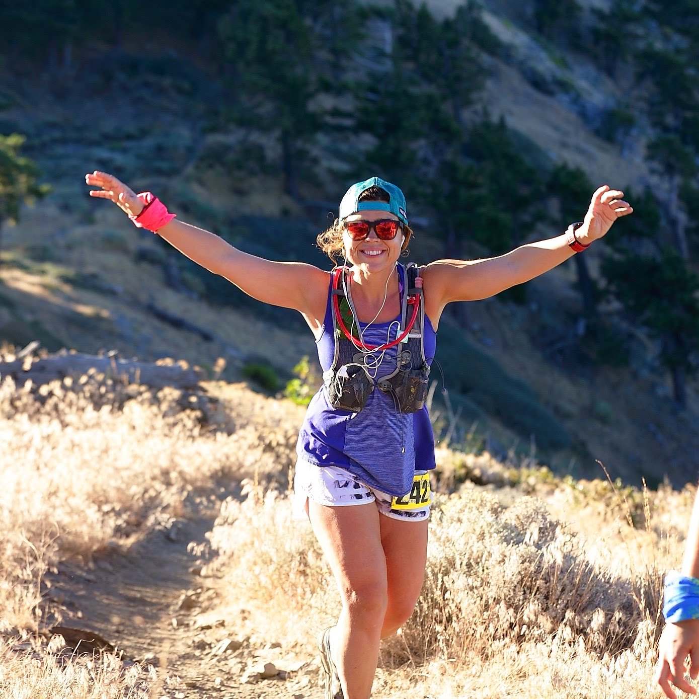 Woman smiling with arms outstretched, wearing sunglasses, a cap, and athletic clothing, outdoors in a natural setting.