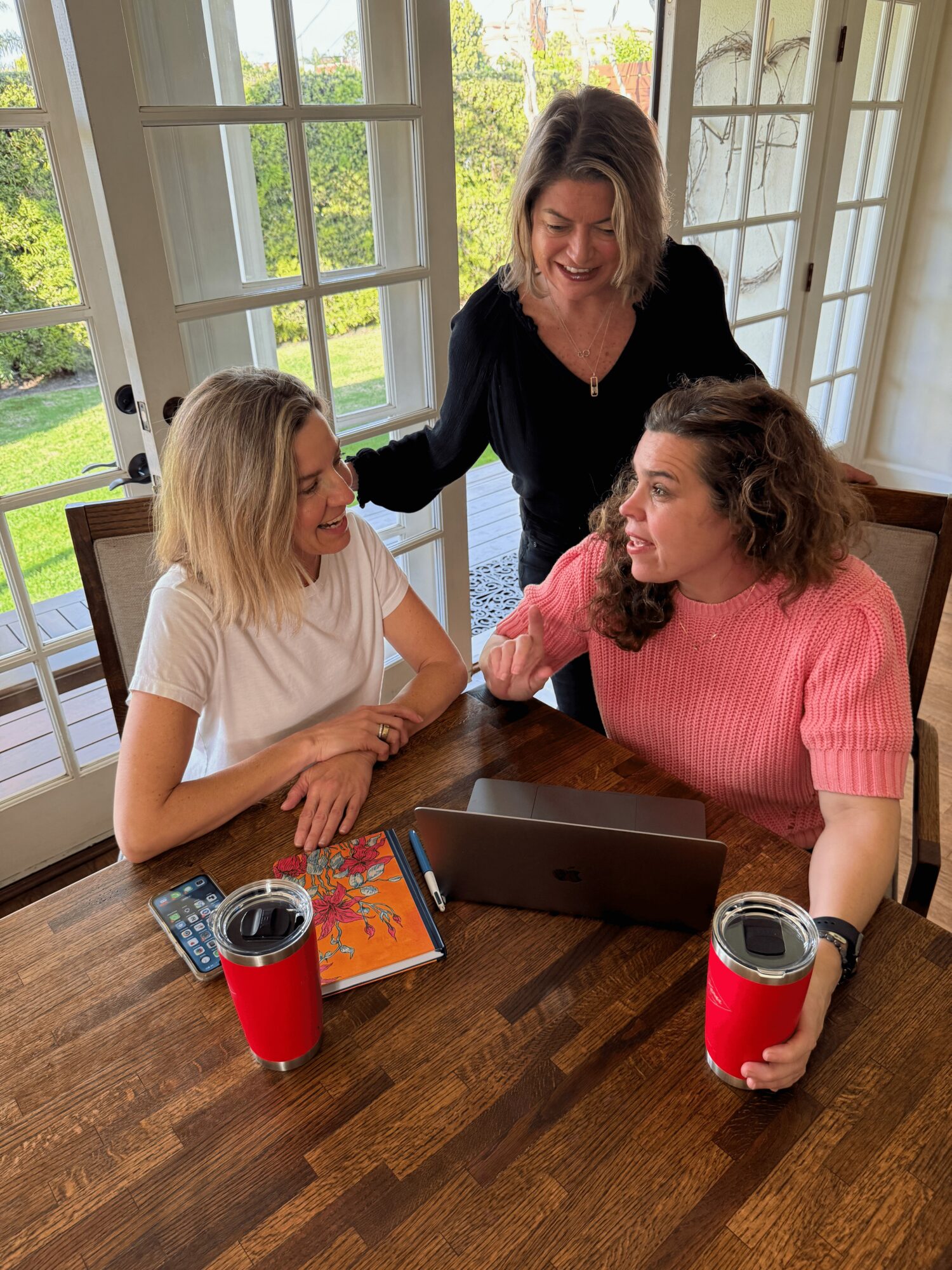 Three women gathered around a table with a laptop, two with drinks and a notebook, smiling indoors near windows.