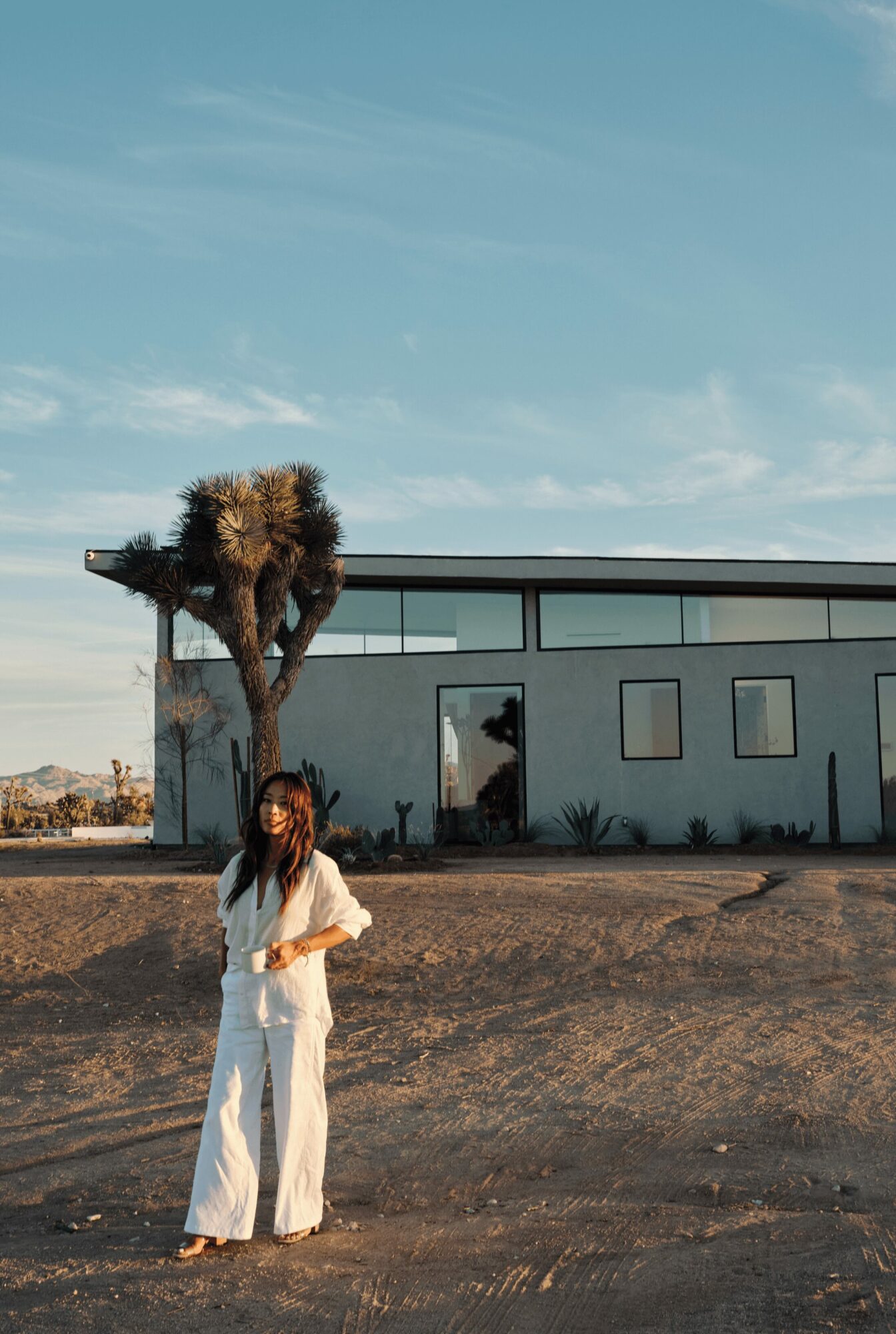 Woman in white pants and top standing outdoors near a tree and modern building under a blue sky.