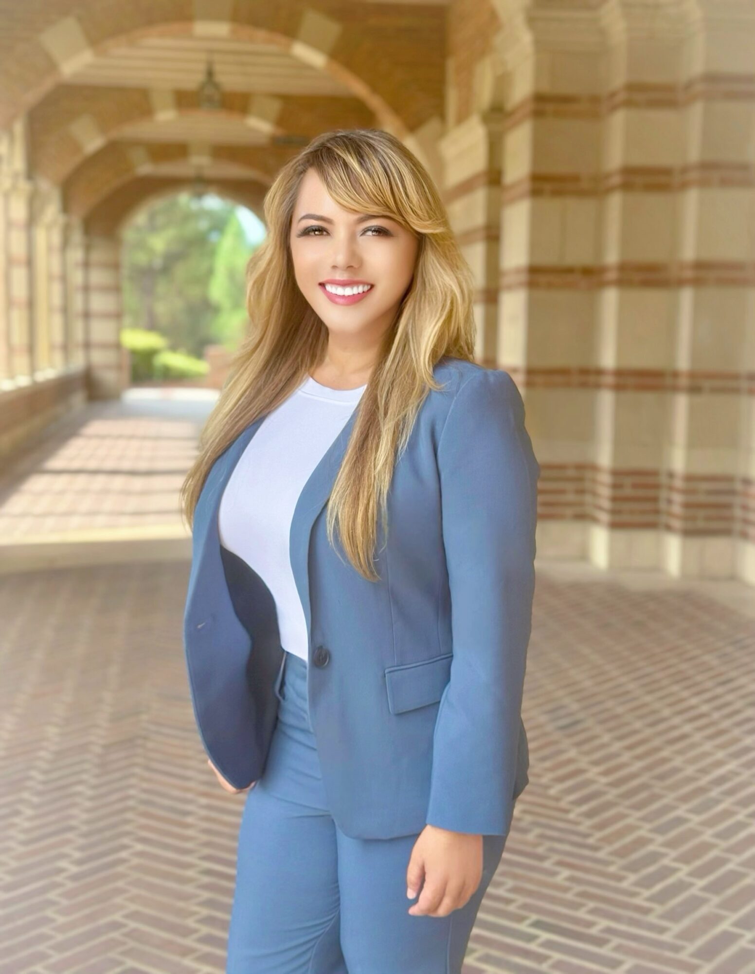 Young woman with long blonde hair smiling, wearing a blue blazer and white shirt, standing outdoors near arches.