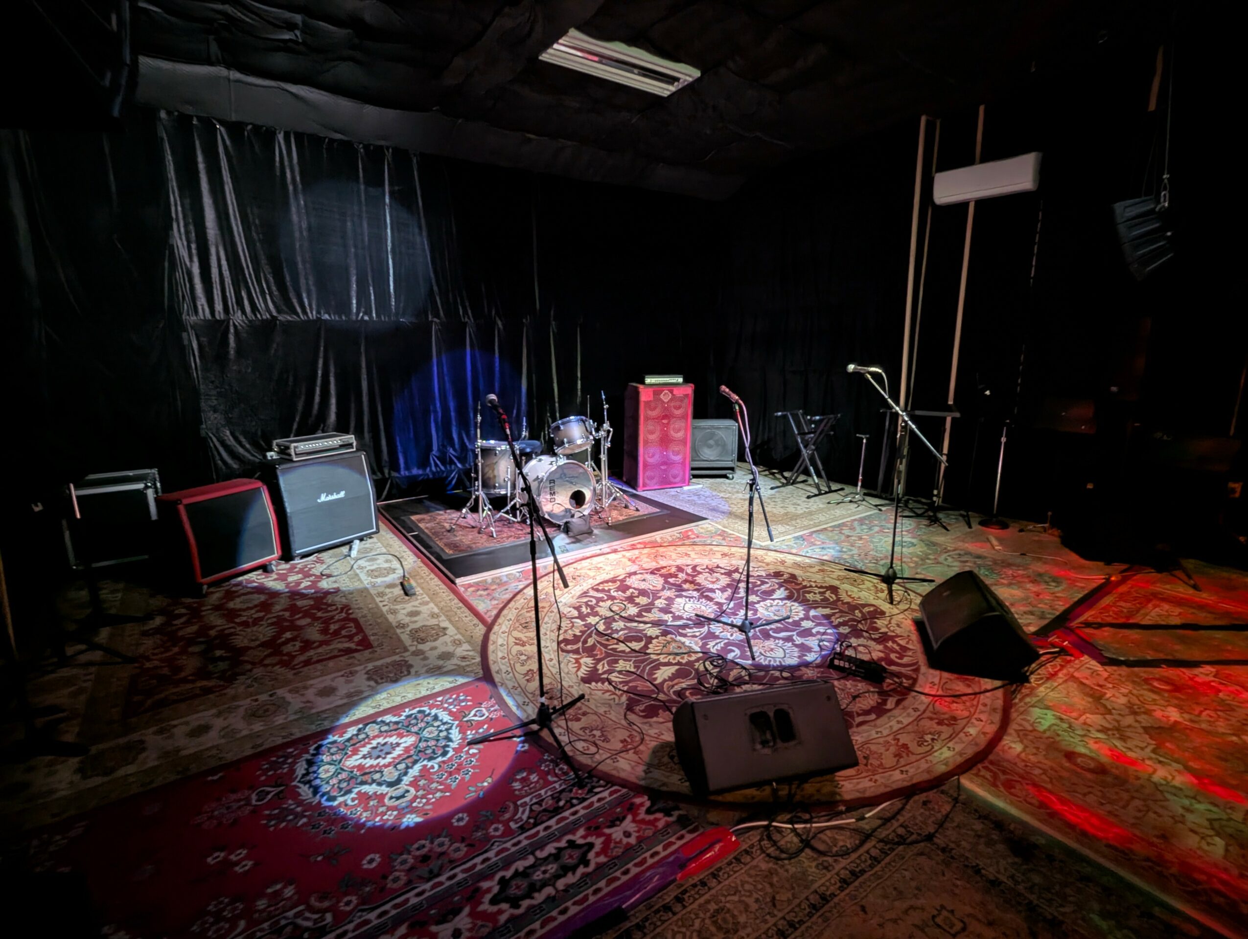 Empty stage with musical instruments, microphones, and amplifiers, set on a decorated carpeted floor with curtains in the background.