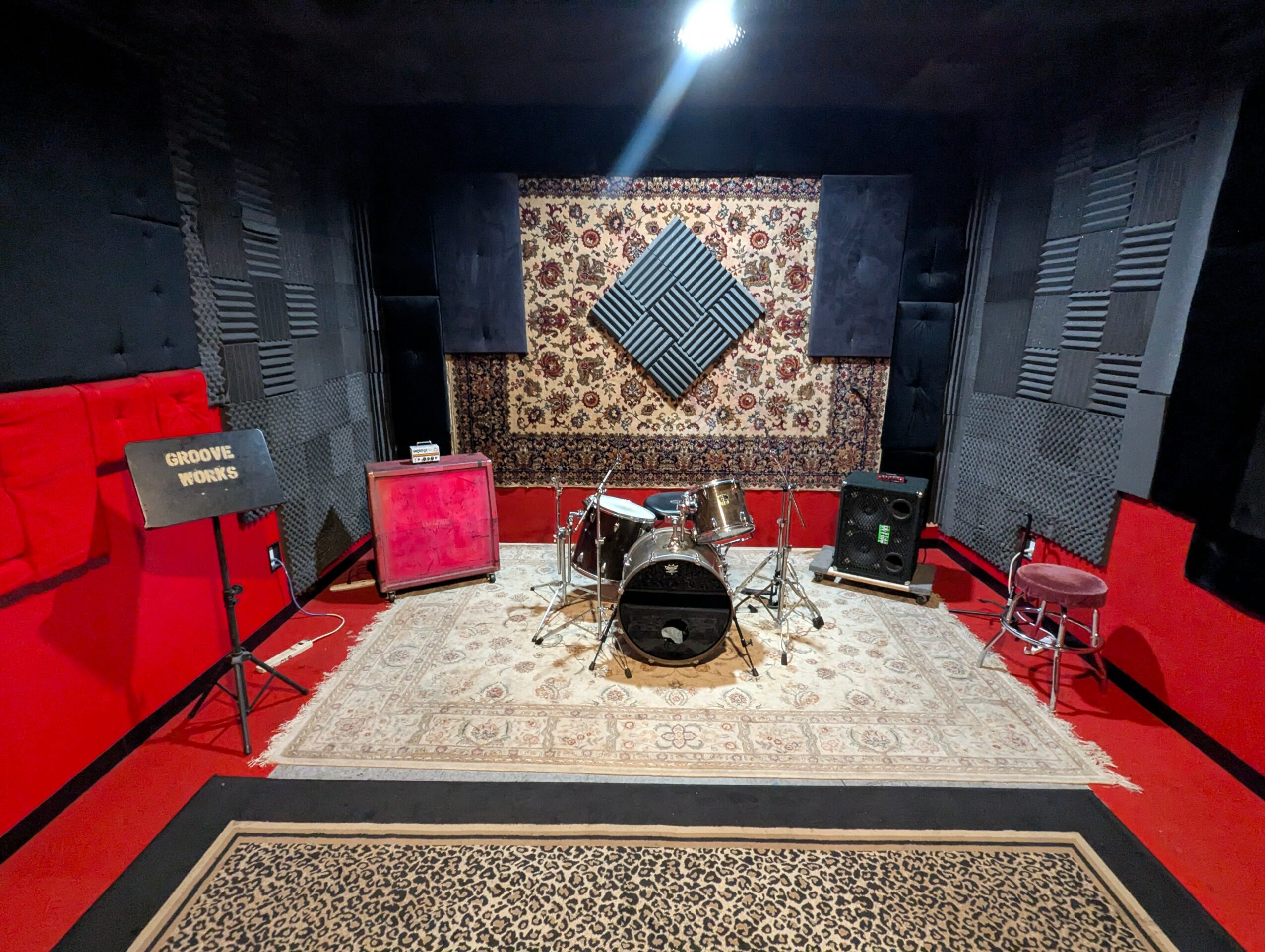 Music rehearsal room with drums, amplifiers, and chairs, decorated with patterned wallpaper and acoustic panels.