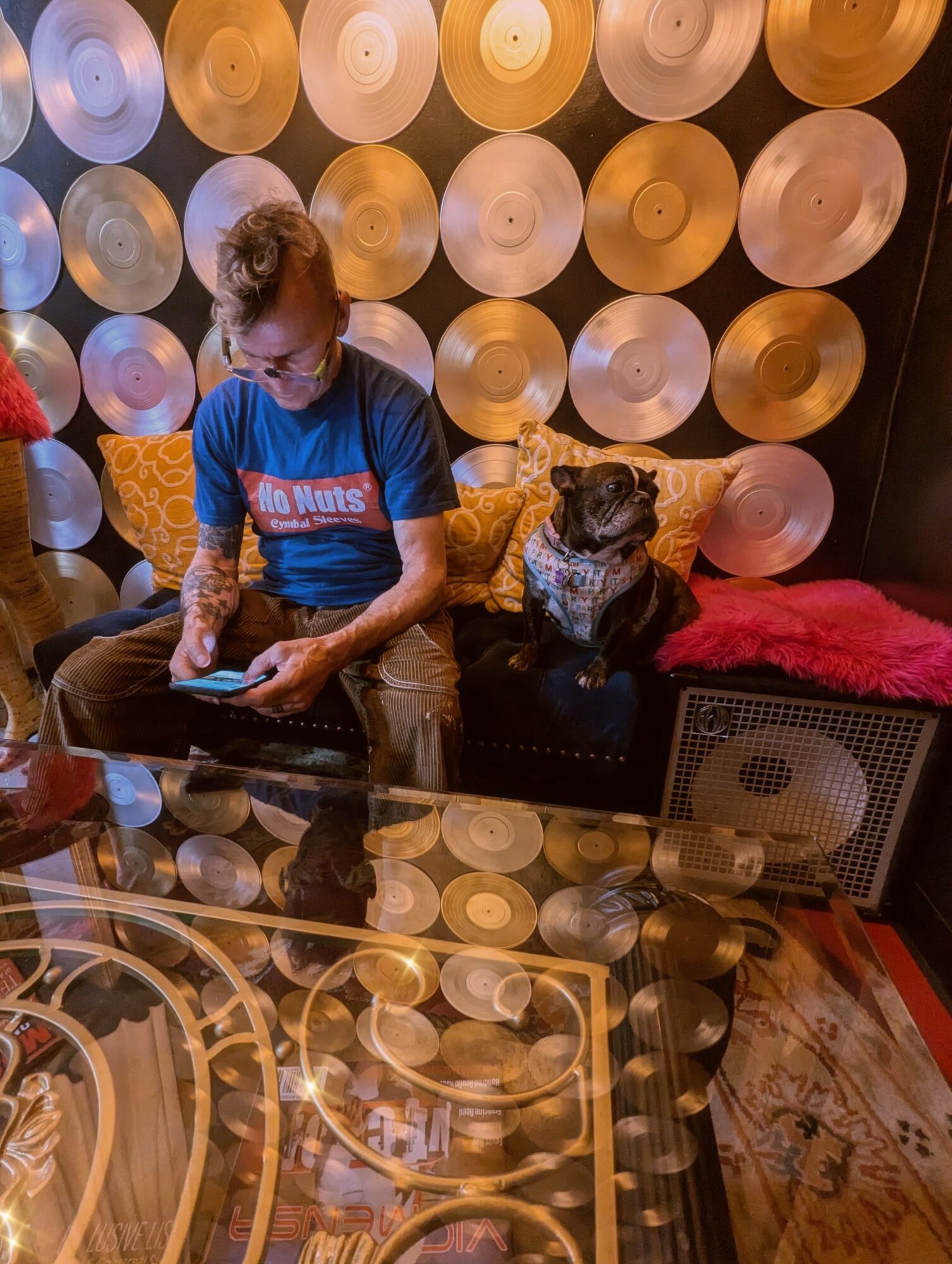 Person with glasses sitting on a black couch with a dog, wall decorated with circular objects, glass table in foreground.