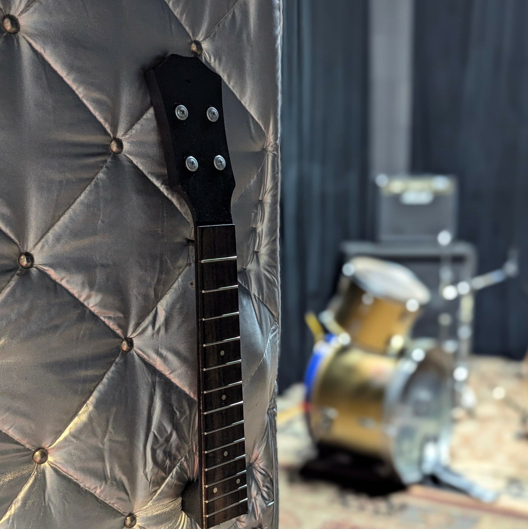 Close-up of a guitar neck leaning against a padded surface with a drum set in the background.